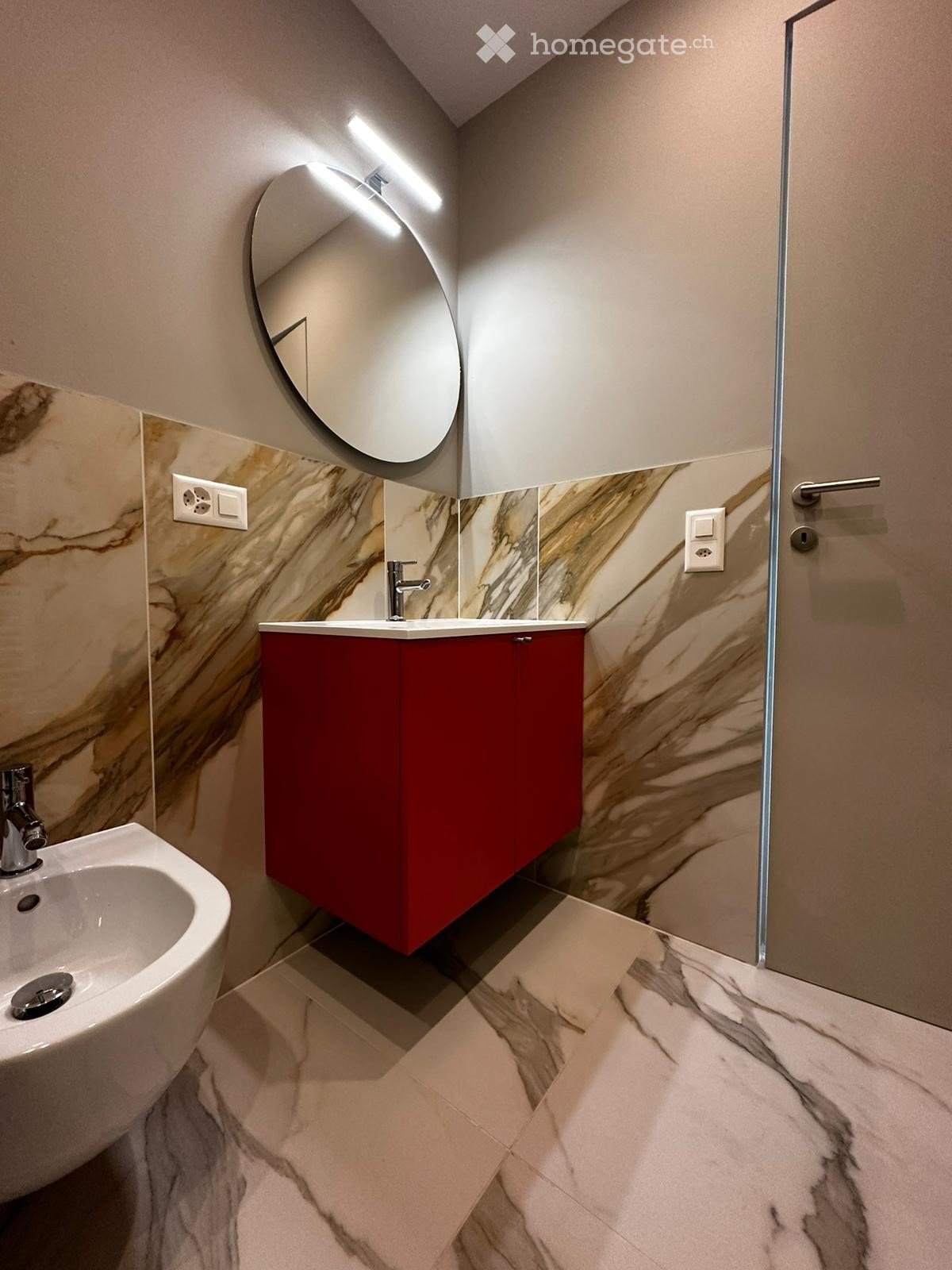 Modern bathroom with marble walls and floor, red cabinet, white sink, and a round mirror with built-in lighting.
