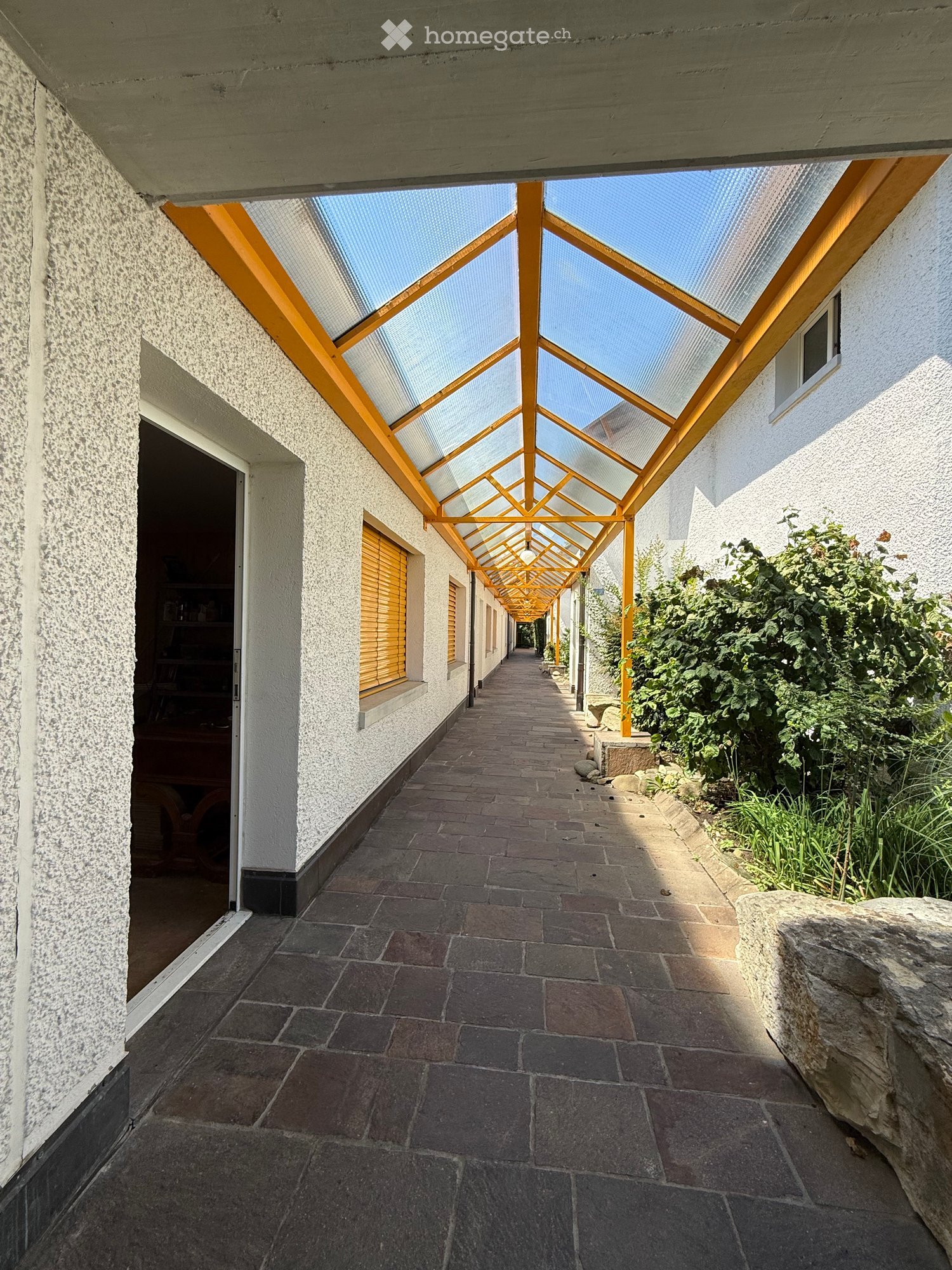 A covered outdoor corridor with a tiled floor, white walls, and a wooden-framed glass roof supported by orange beams. The corridor is flanked by greenery and has windows with wooden shutters on one side.