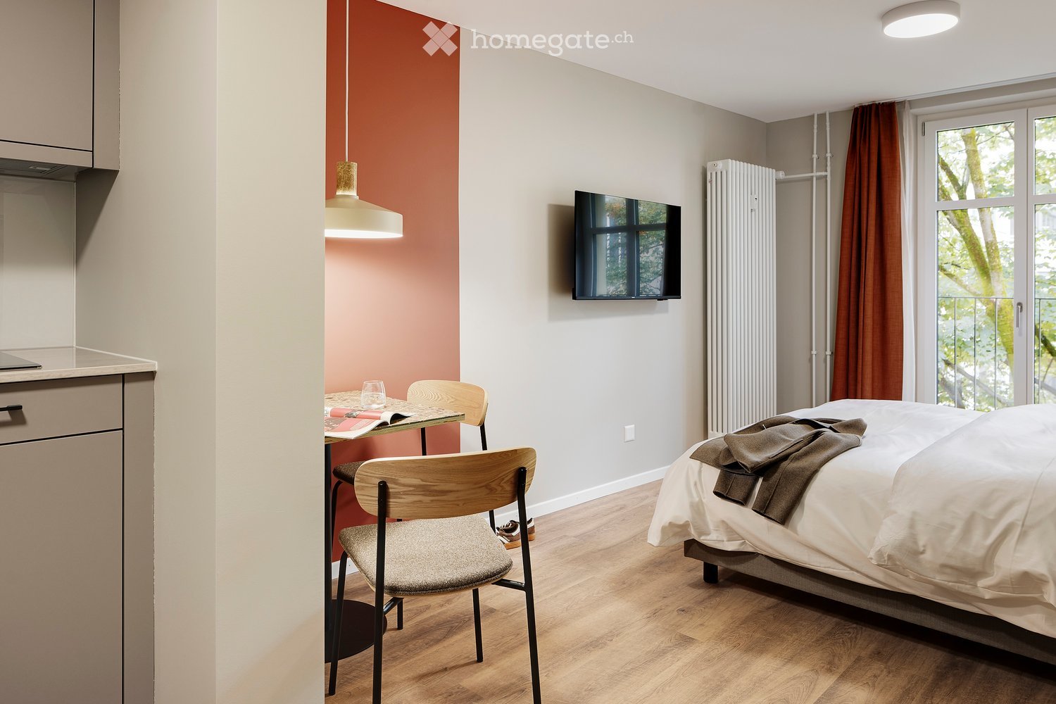 Modern, compact bedroom with a bed, a small table with chairs, a television mounted on the wall, and a curtained window. The room features a pendant light and a radiator.