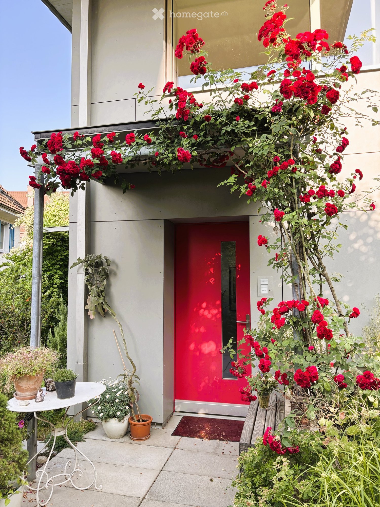 Garden area with a red front door, white metal table, potted plants, and climbing roses on the wall.