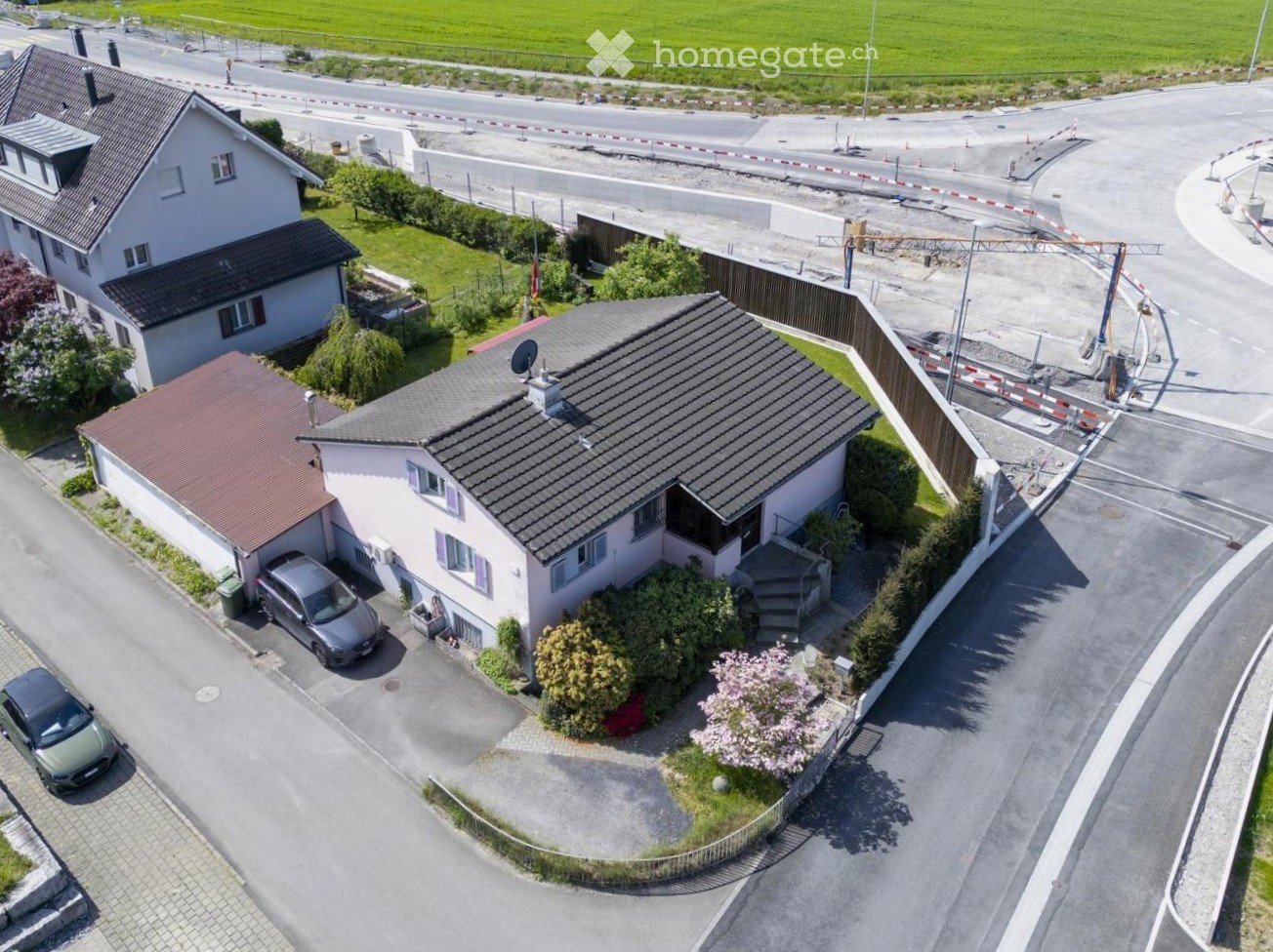 Aerial view of a two-story house with a tiled roof, surrounded by greenery and a driveway with parked cars.