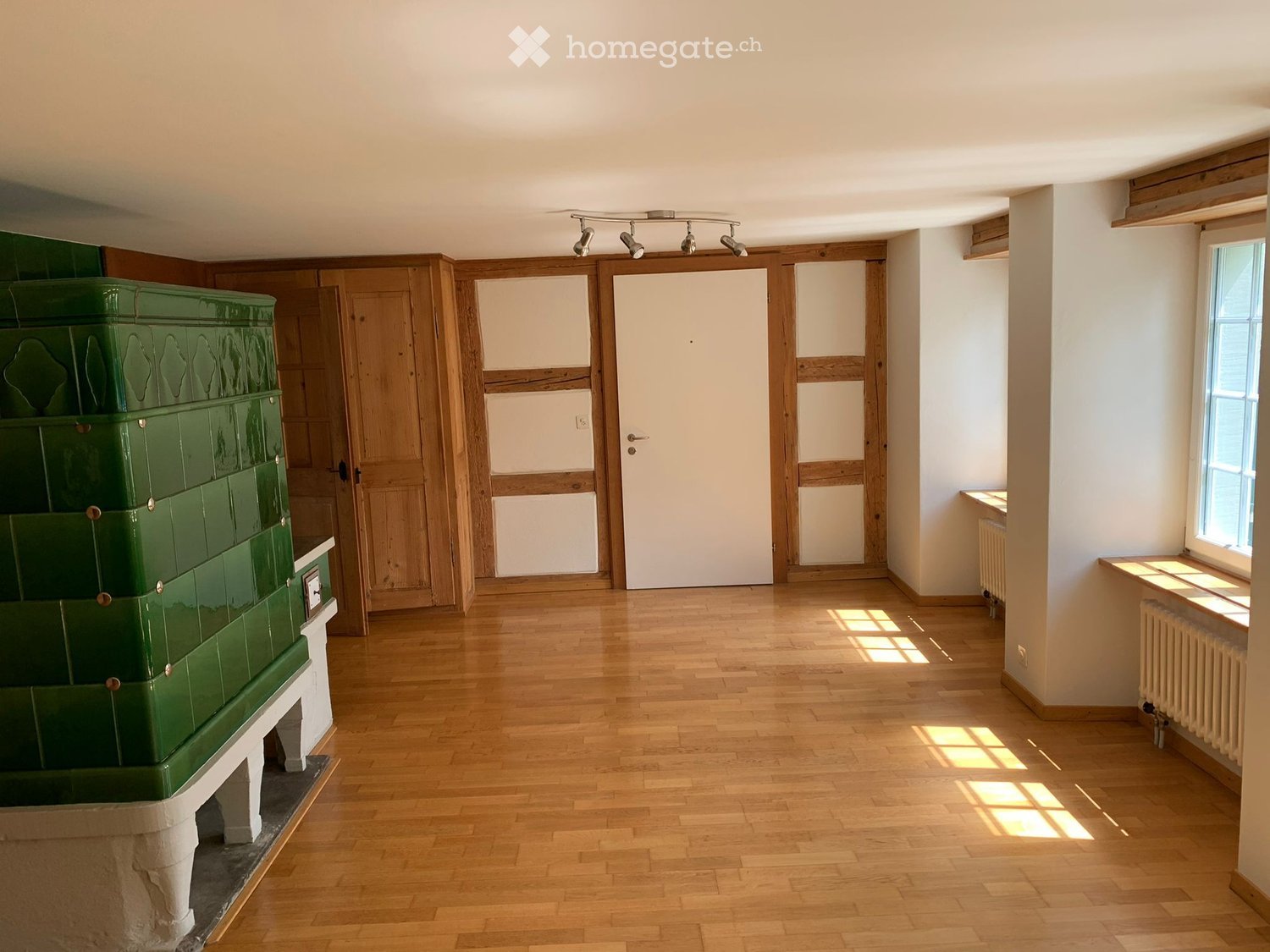 Empty room with wooden flooring, white walls, and a large green tiled stove. Natural light streams in from a window on the right side.