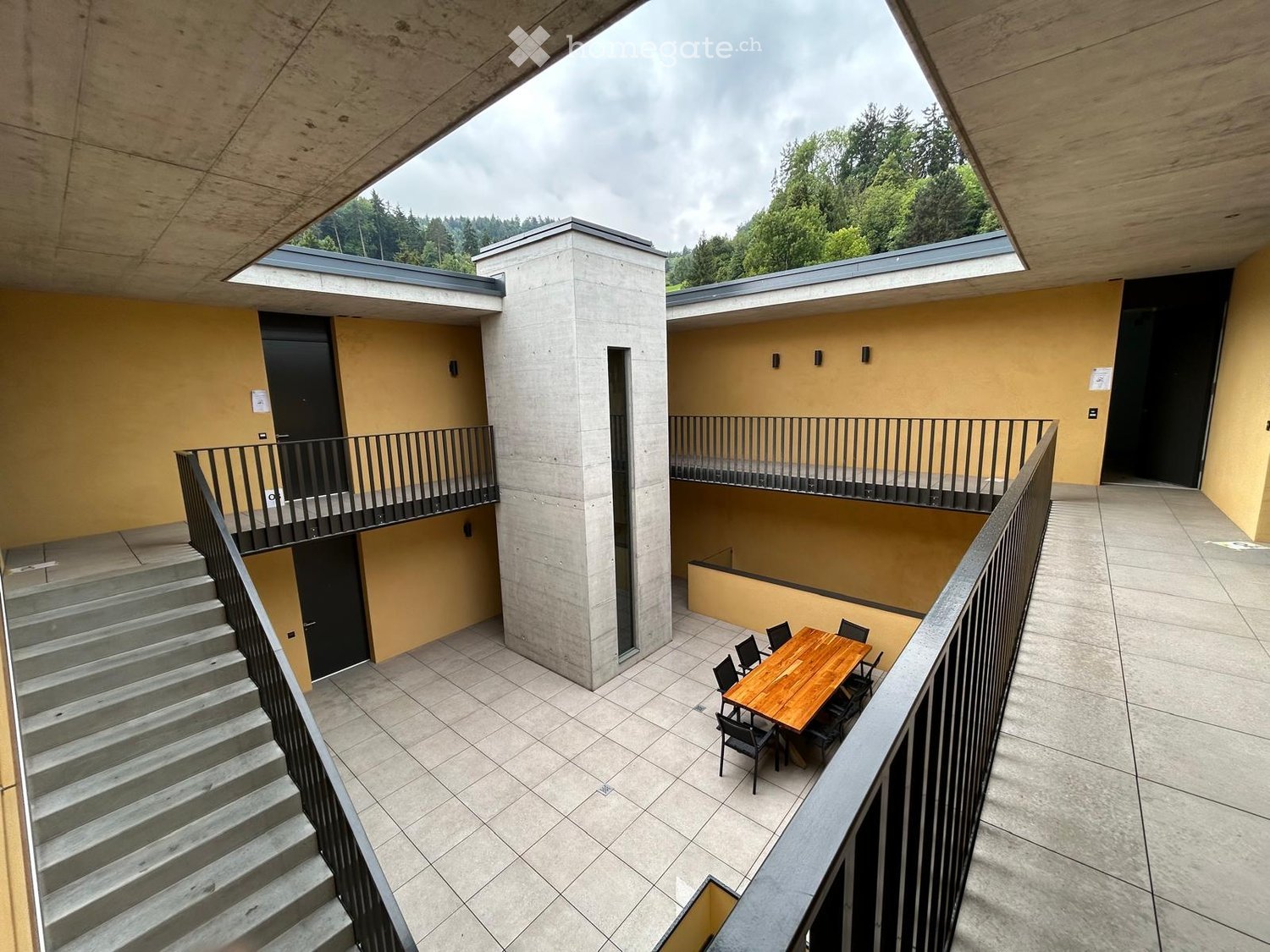 Modern balcony with tiled floor, wooden table, and black metal chairs, overlooking a landscaped area with trees.