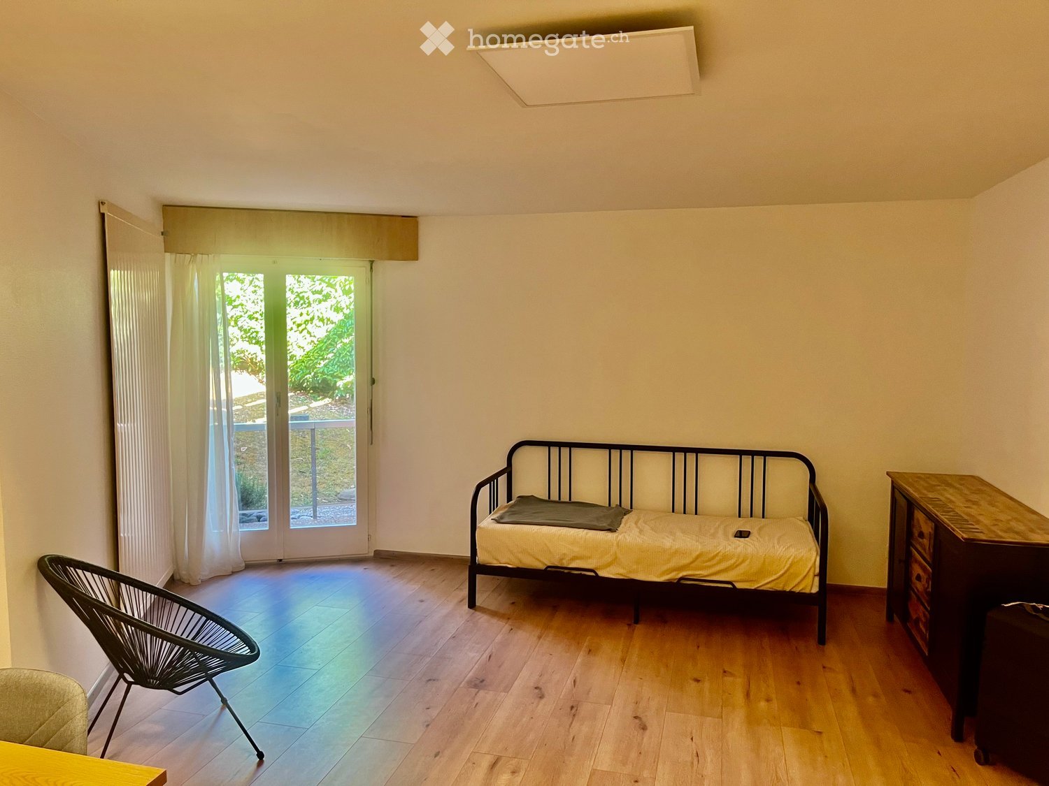A small, furnished room with a wooden floor, a sofa, a chair, a small table, and a cabinet. The room has large windows with curtains and a door leading to a balcony.