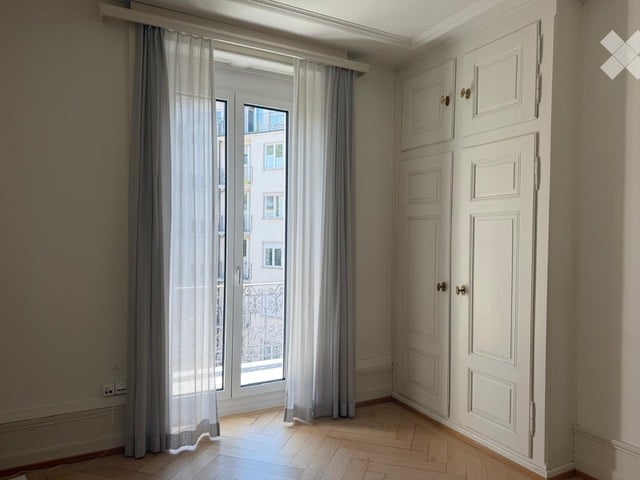 Empty room with wooden flooring, large window with grey curtains, white built-in cabinetry, and a view of a building outside.