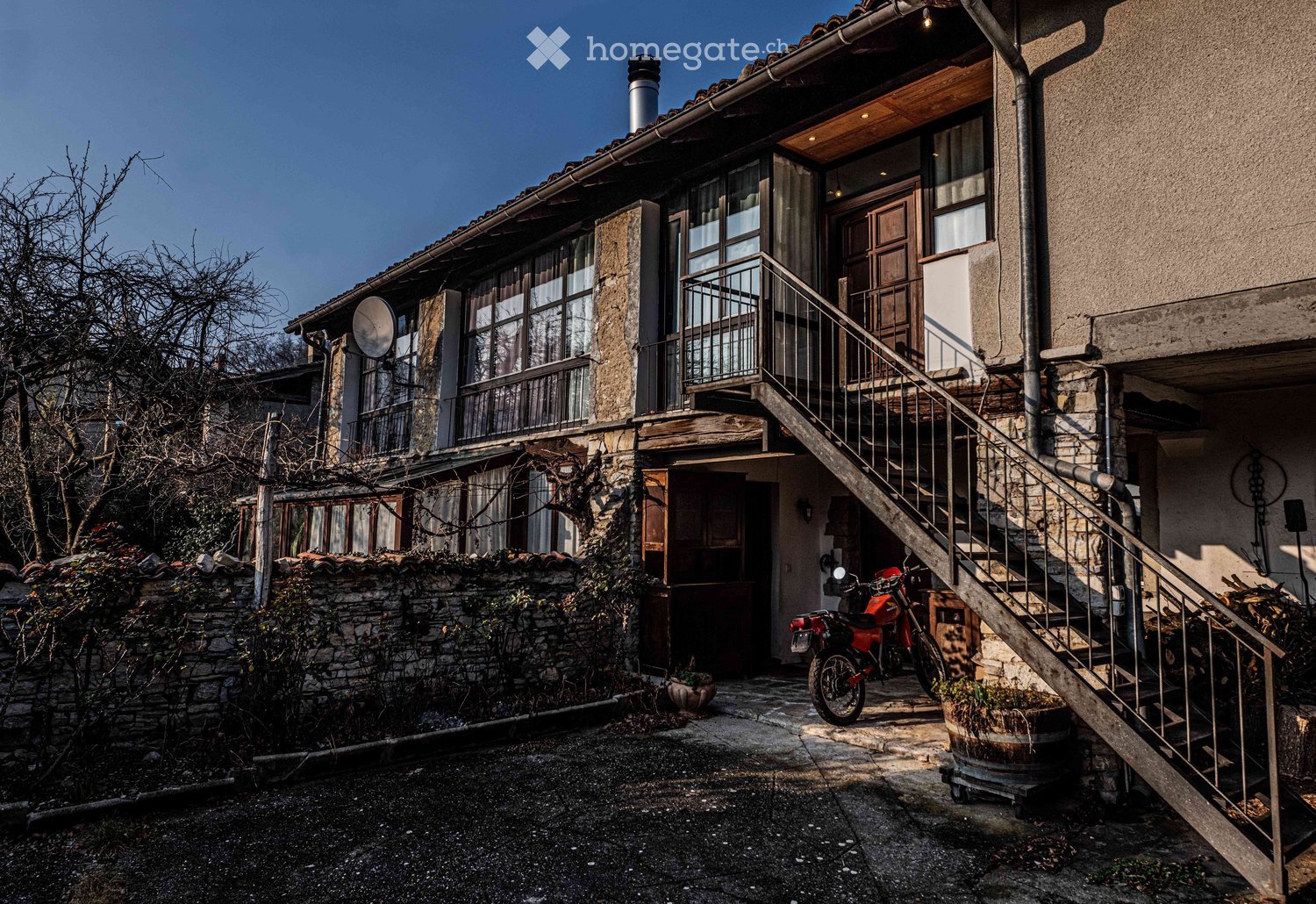 Old house with stone walls, metal staircase, motorcycle parked, satellite dish, and a barrel.