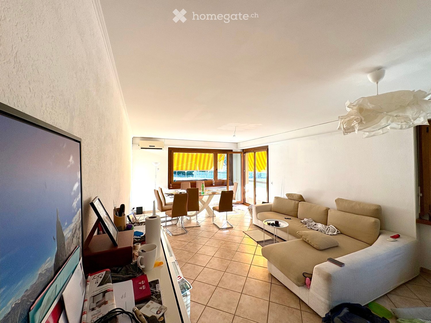 Living room with a beige sofa, coffee table, dining area with chairs and table, large screen, and a view to a balcony.