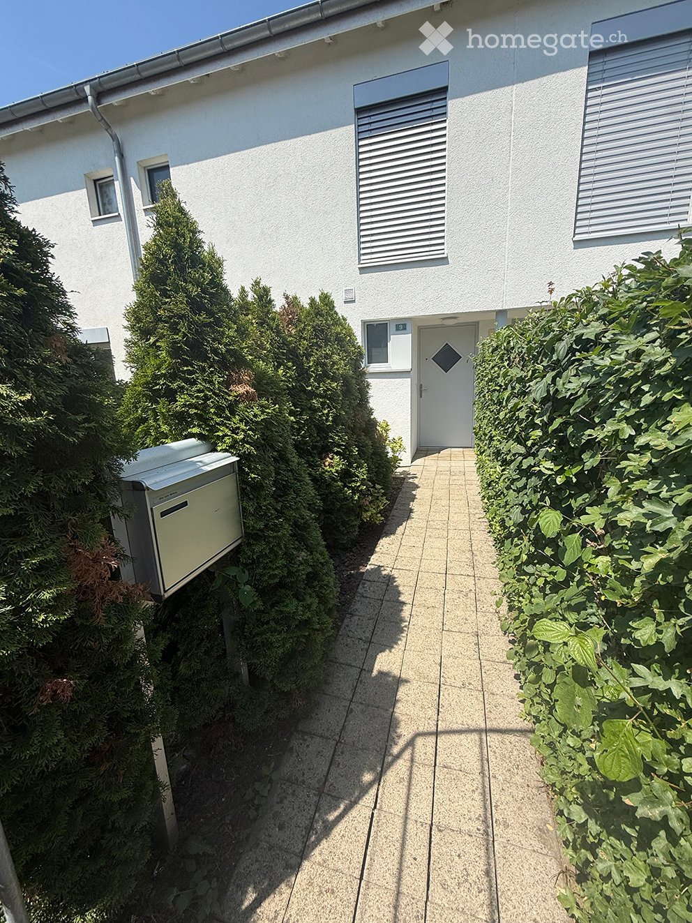 A pathway leads to a white building entrance, flanked by neatly trimmed hedges and a small utility box on the left.