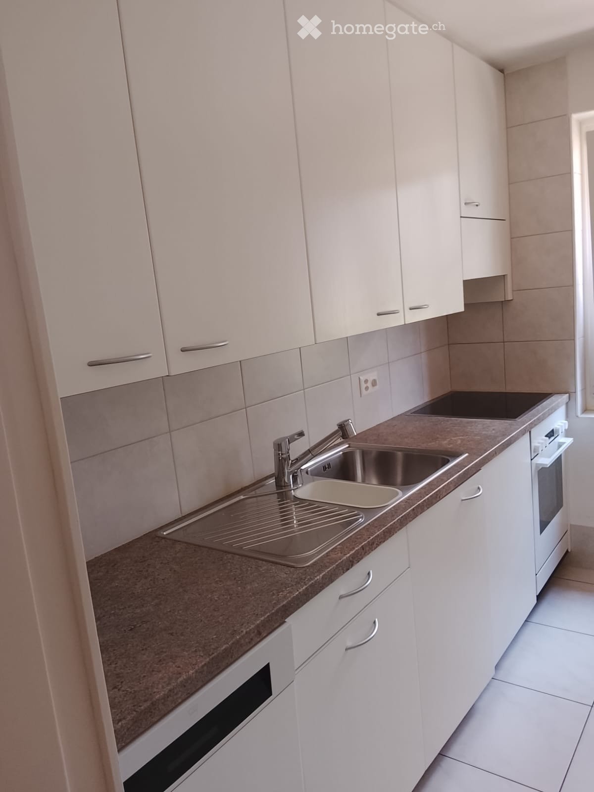 Modern kitchen with white cabinets, a stainless steel sink, and built-in oven. Countertop is brown with a tiled backsplash.