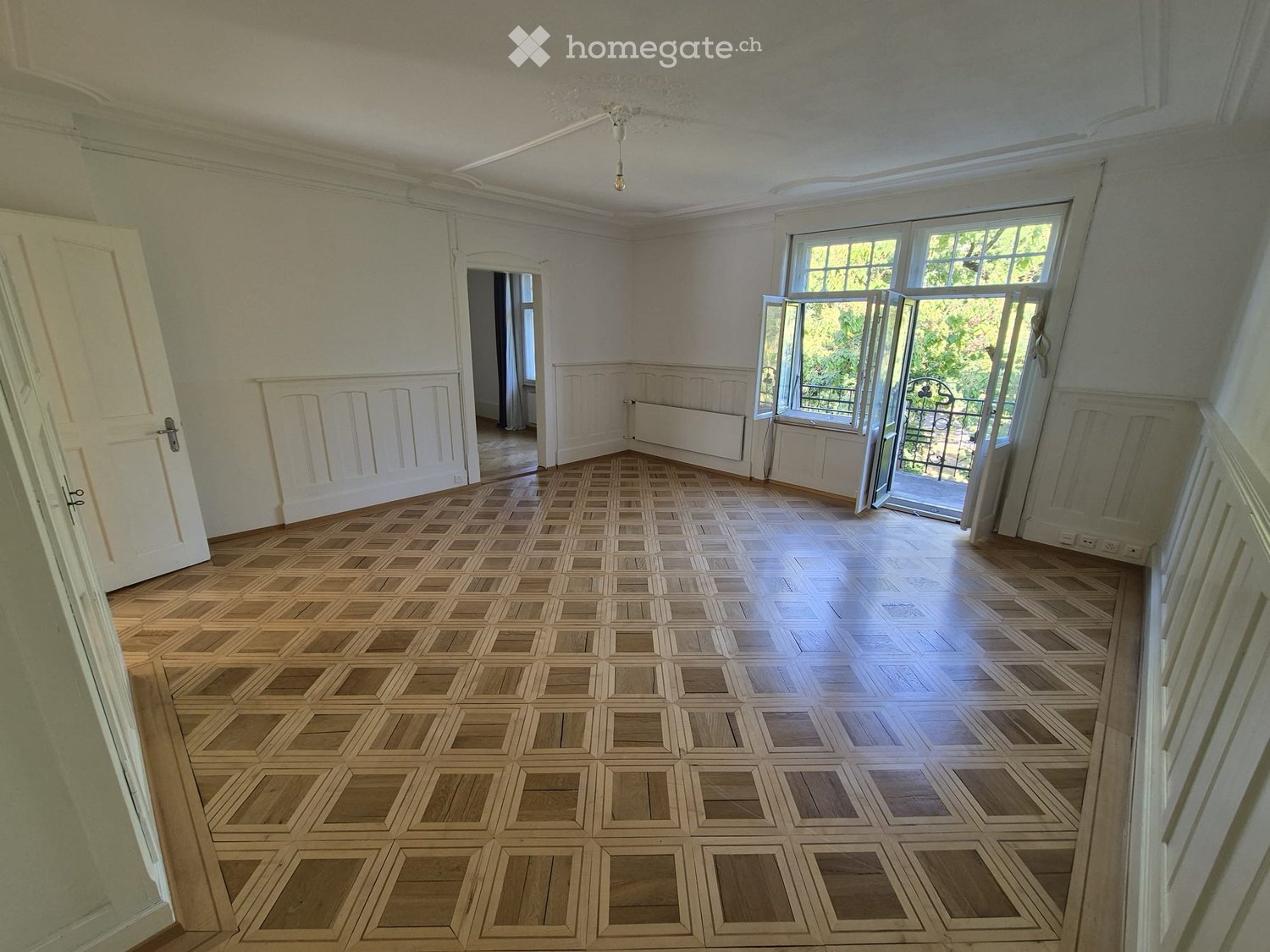 Empty room with wooden parquet flooring, white walls, large windows, and a visible radiator. No furniture or equipment present.