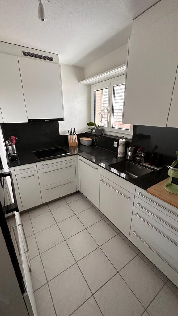 Modern kitchen with white cabinets, black countertops, and tiled flooring. Features include a sink, faucet, blender, bowl, knife block, toaster, cup, bottle, and fridge.