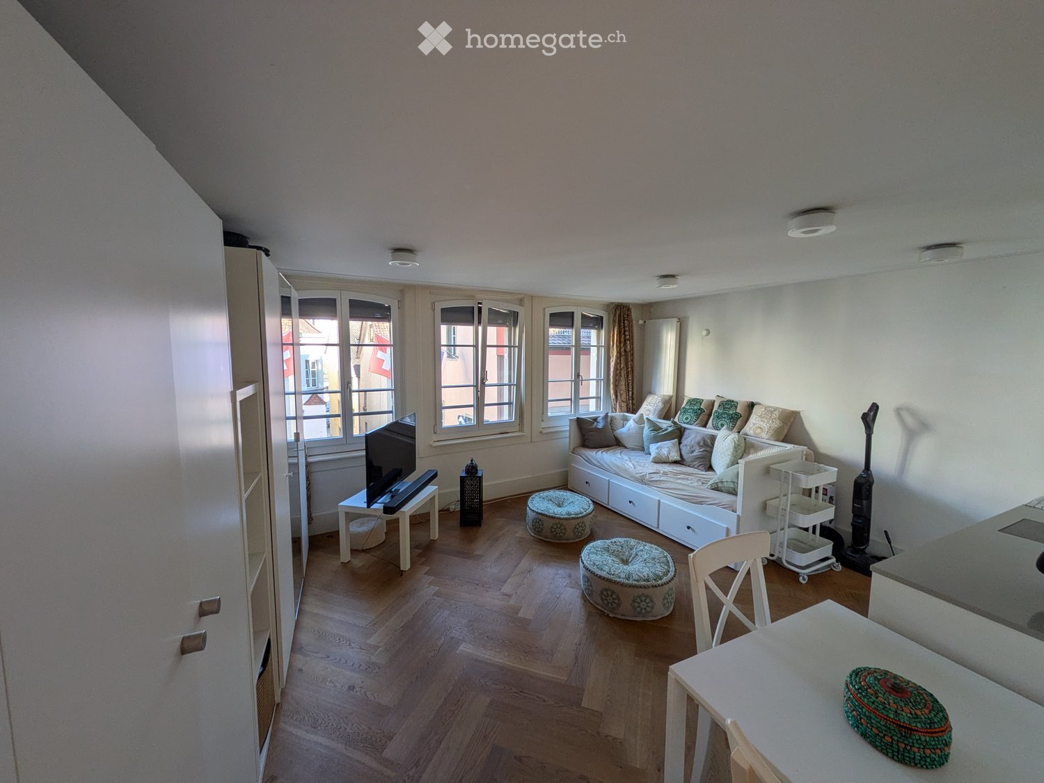 A modern, well-lit living space with wooden flooring, large windows, a sofa, a TV, and a small dining area.