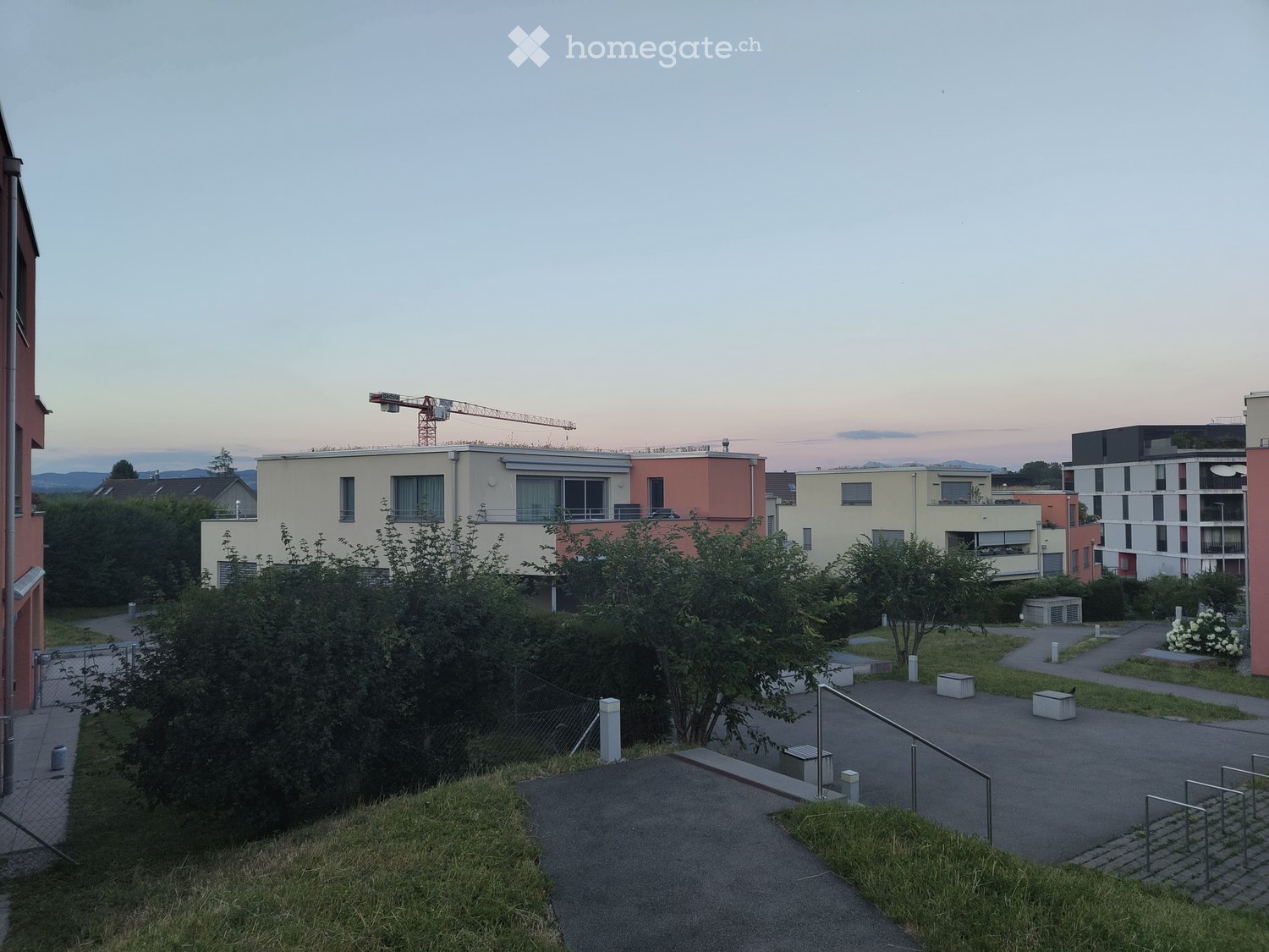 View of a residential area with multiple buildings, greenery, and a crane in the background.