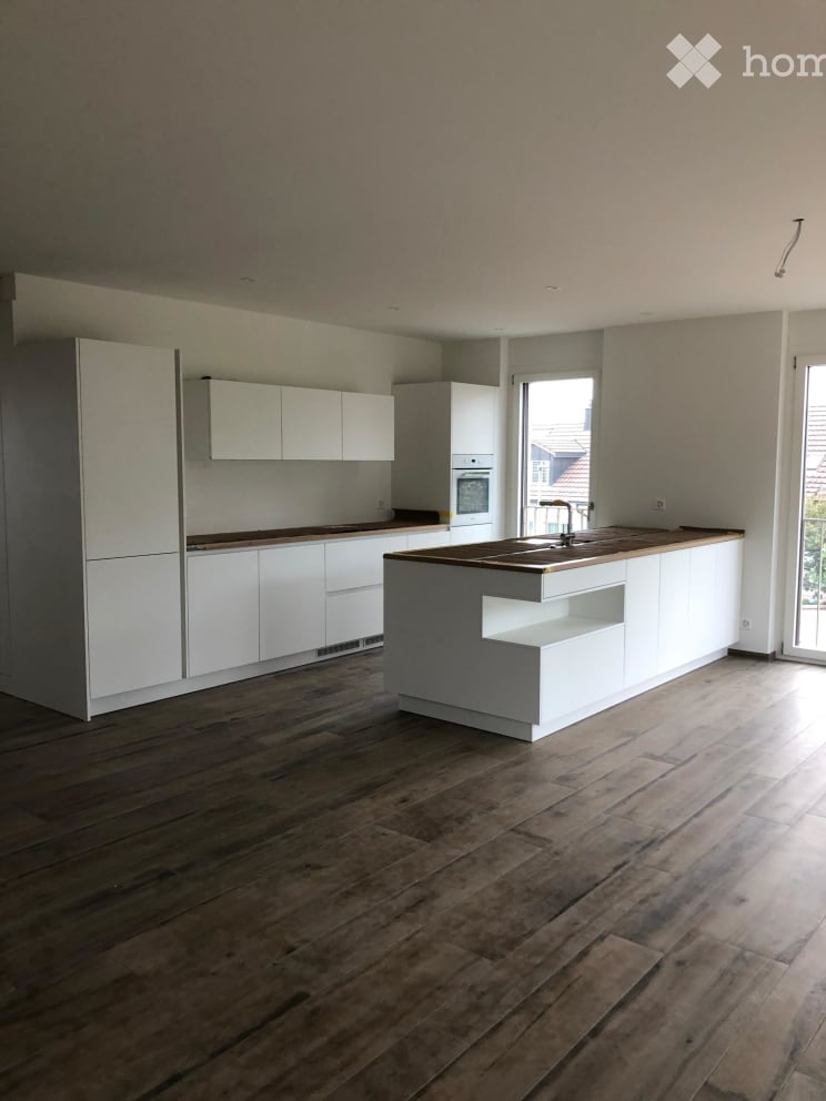 Modern kitchen with white cabinets, wooden countertops, and a central island. The floor is made of dark wood planks, and the space is well-lit with natural light from a large window.