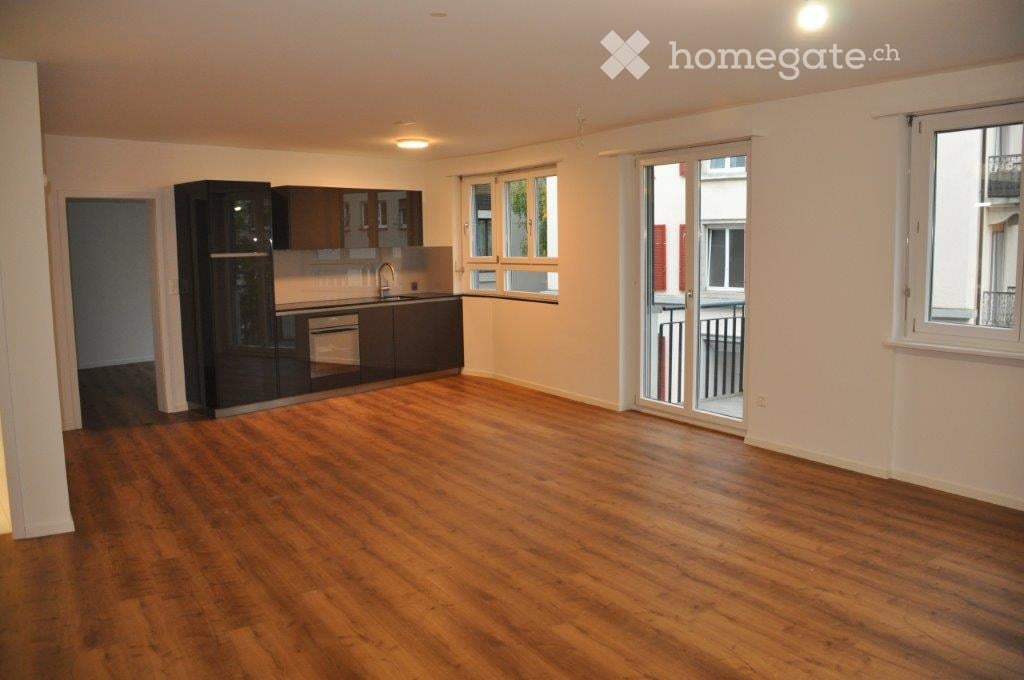 Empty room with wooden flooring, white walls, and a modern kitchen area with cabinets and a sink. Features large windows and a sliding glass door leading to a balcony.
