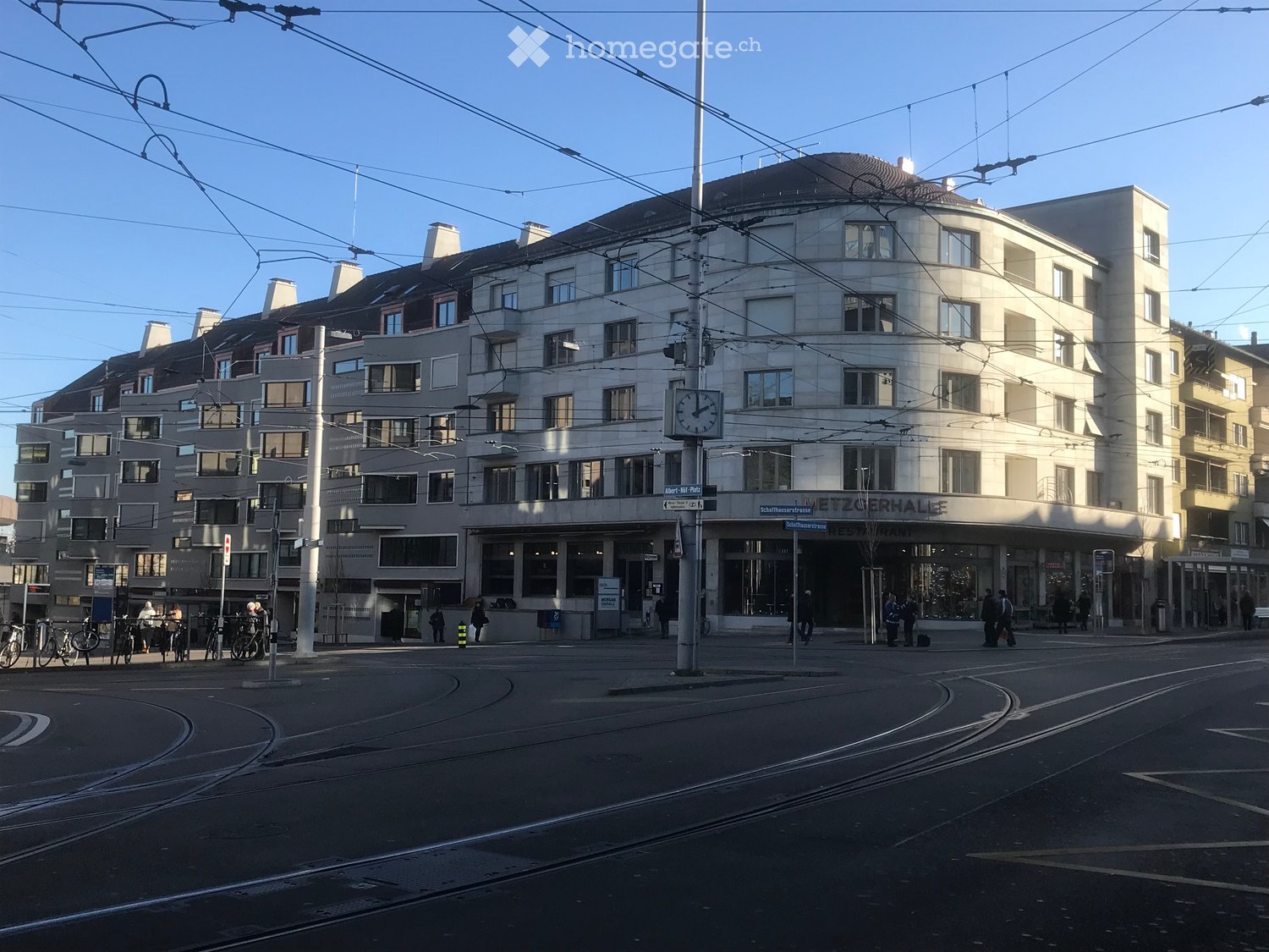 Multi-story building with a curved facade, large windows, and a prominent entrance labeled 'Schlafhausrestaurant'. The building is surrounded by a street with tram tracks, a clock, and several people walking.