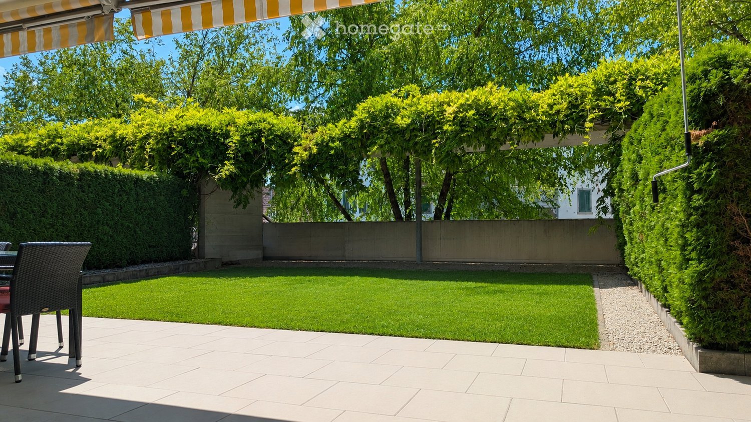 Well-maintained garden with a neatly trimmed lawn, surrounded by hedges and trees, featuring a patio area with a wicker chair and table under a striped awning.