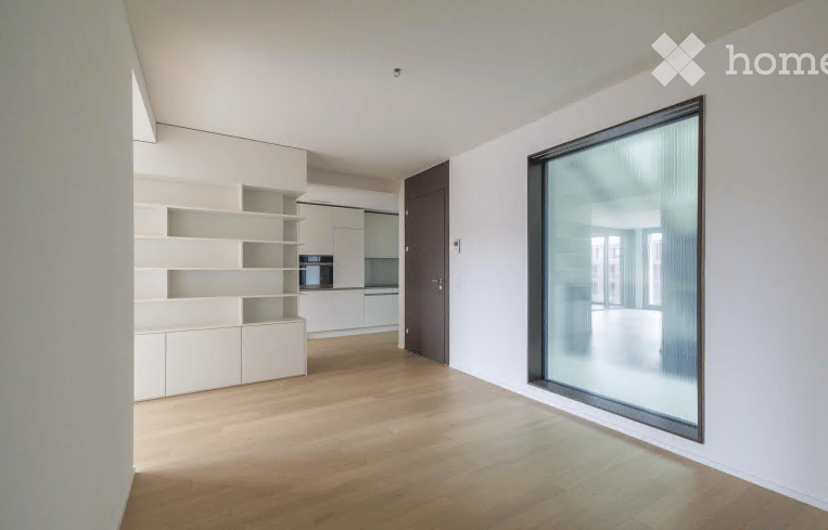 Modern, minimalist room with light wooden flooring, white walls, built-in shelves, and a small kitchen area featuring a microwave and cabinets.