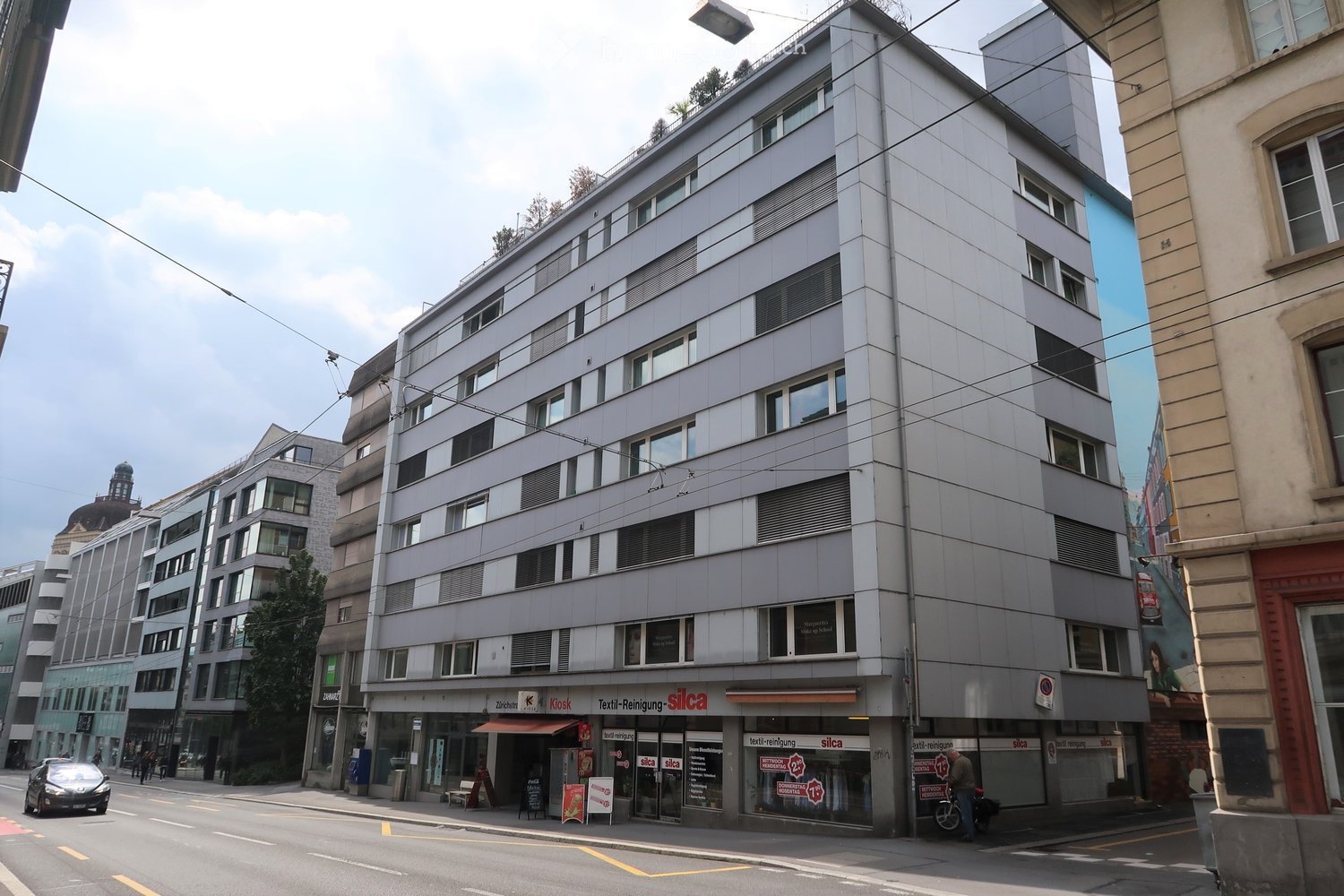 Modern multi-story building with grey facade, large windows, and commercial storefronts on the ground floor, including a textile cleaning shop.