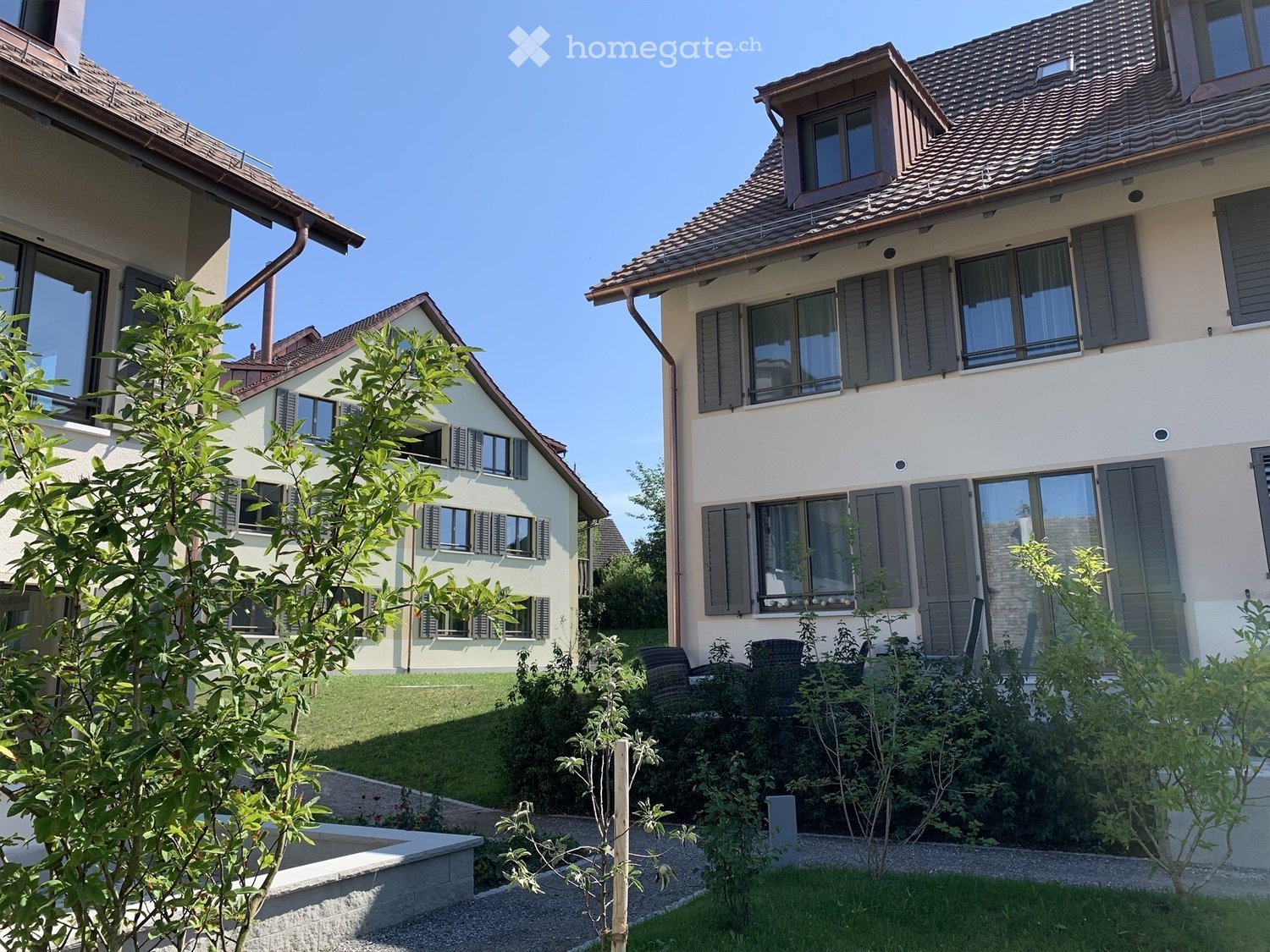 A well-maintained garden area with a gravel pathway, small trees, shrubs, and a lawn, situated between two modern houses with light-colored facades and dark shutters.