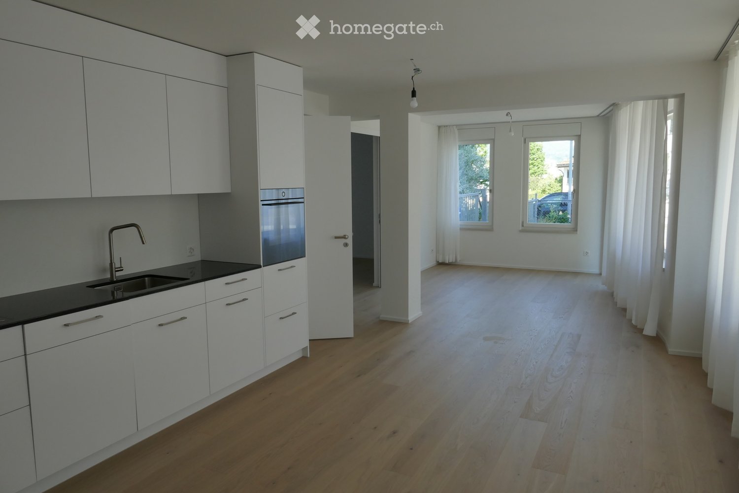 Bright, unfurnished room with light wooden flooring, white walls, and large windows with white curtains. The adjacent kitchen features white cabinets, a black countertop, and a stainless steel sink.
