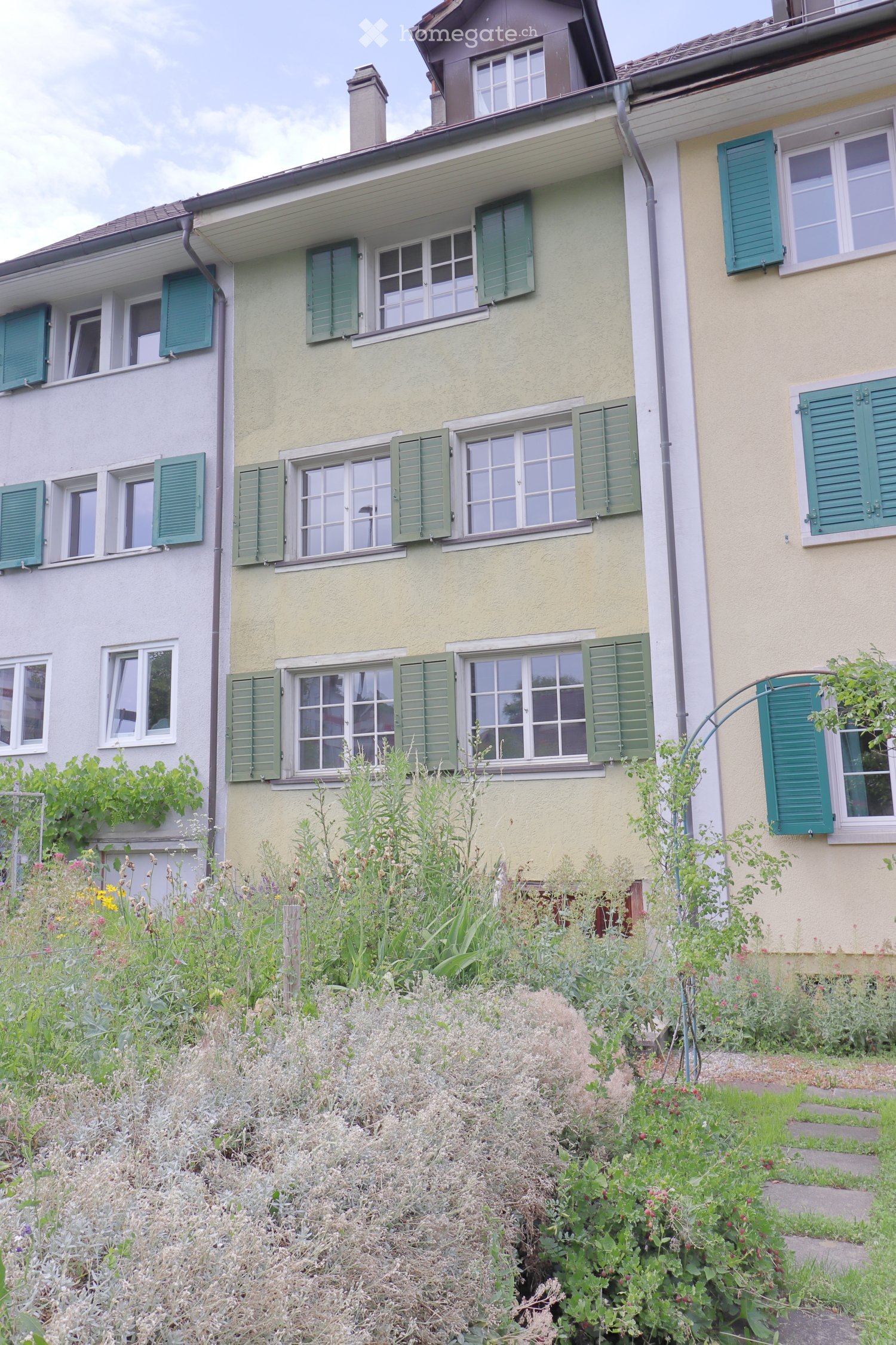 Two-story house with green shutters and multiple windows, surrounded by a garden with various plants and flowers.