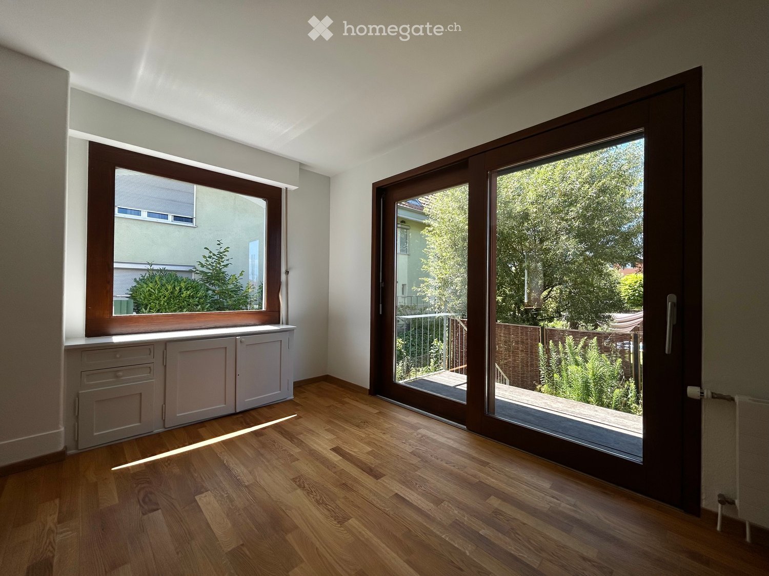 Empty room with wooden flooring, large sliding glass door leading to a balcony, and a window with blinds.