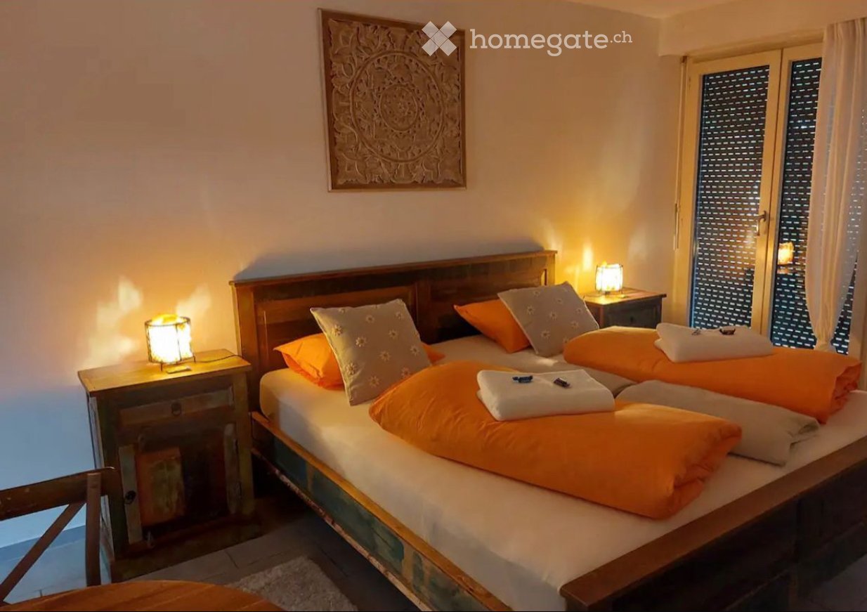 Cozy bedroom with two single beds, orange and white pillows, nightstands with lamps, and a decorative wall piece above the beds.