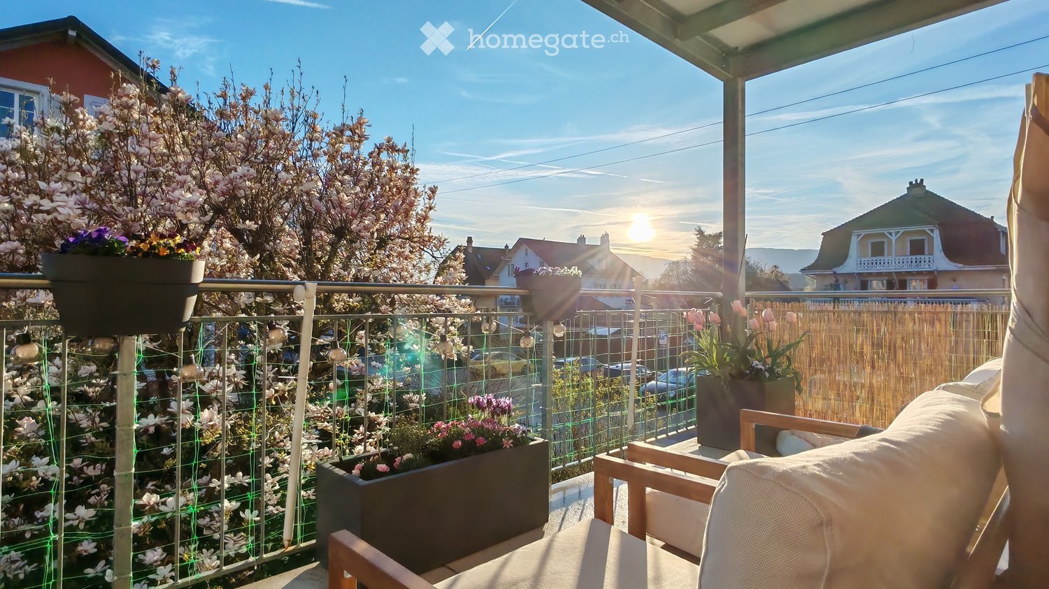 Balcony with wooden chairs, potted plants, and a view of the neighborhood with houses and trees.