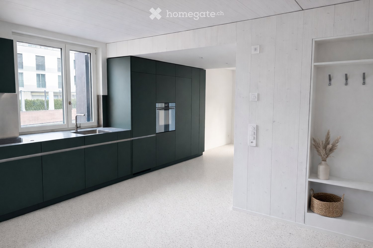 Modern kitchen with dark green cabinets, white wooden walls, and a white tiled floor. Features a sink, faucet, oven, and built-in shelving with decorative items.