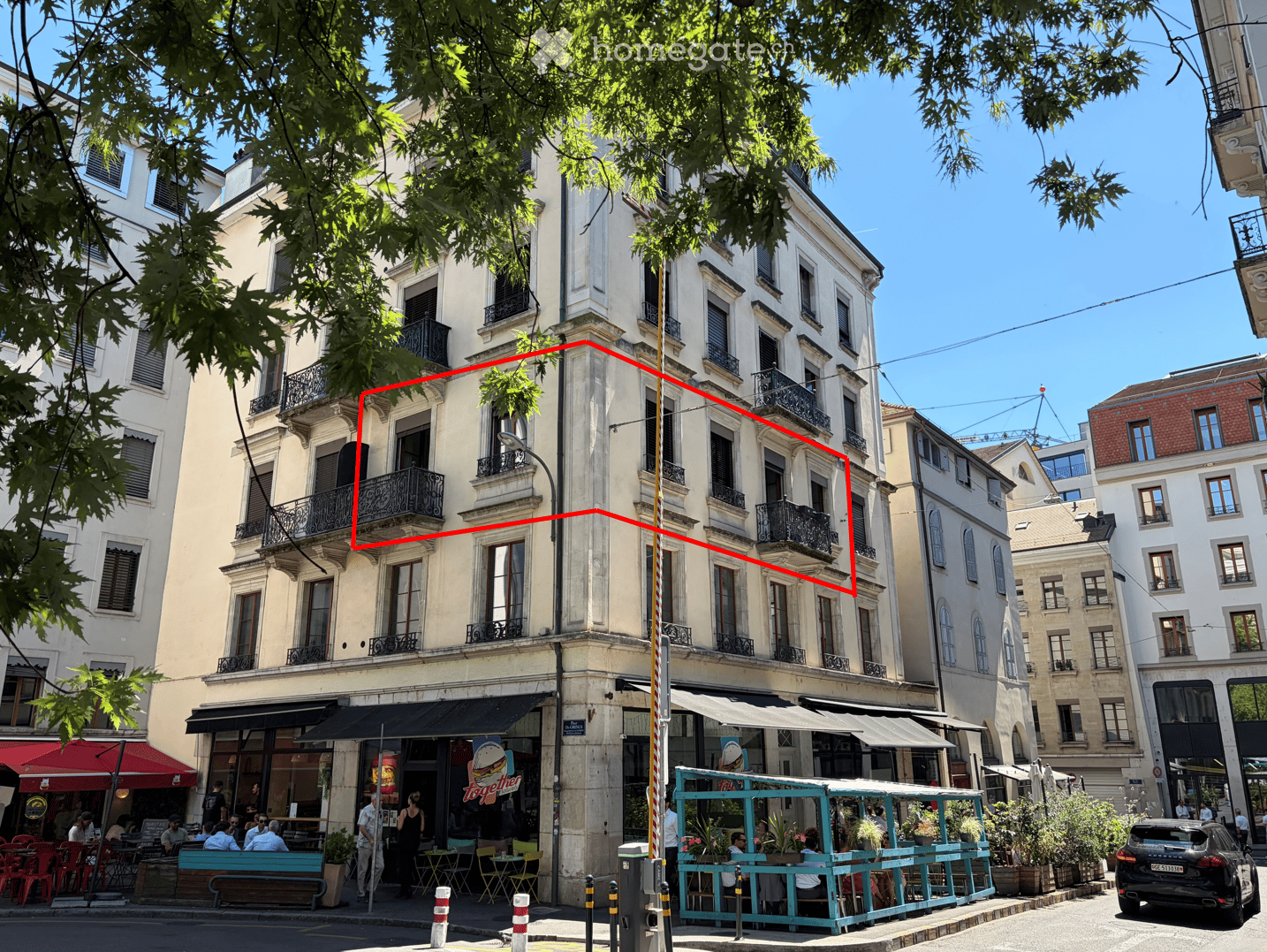 A multi-story building with classic architecture, featuring balconies and large windows. The ground floor has a restaurant with outdoor seating, and the street is lined with trees and other buildings.