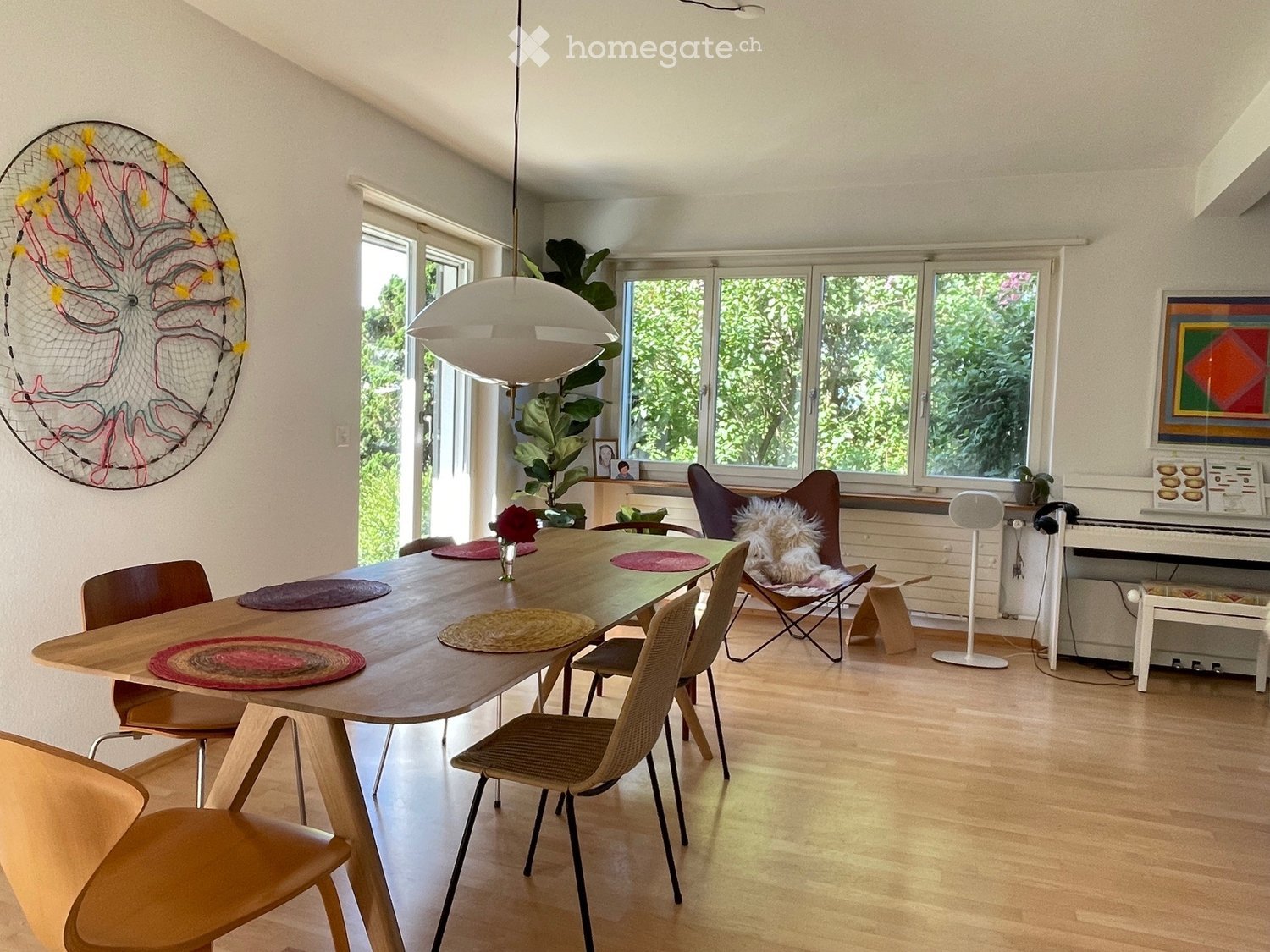 Modern dining room with a large wooden table, multiple chairs, a hanging lamp, a piano, and a large window with a view of greenery.