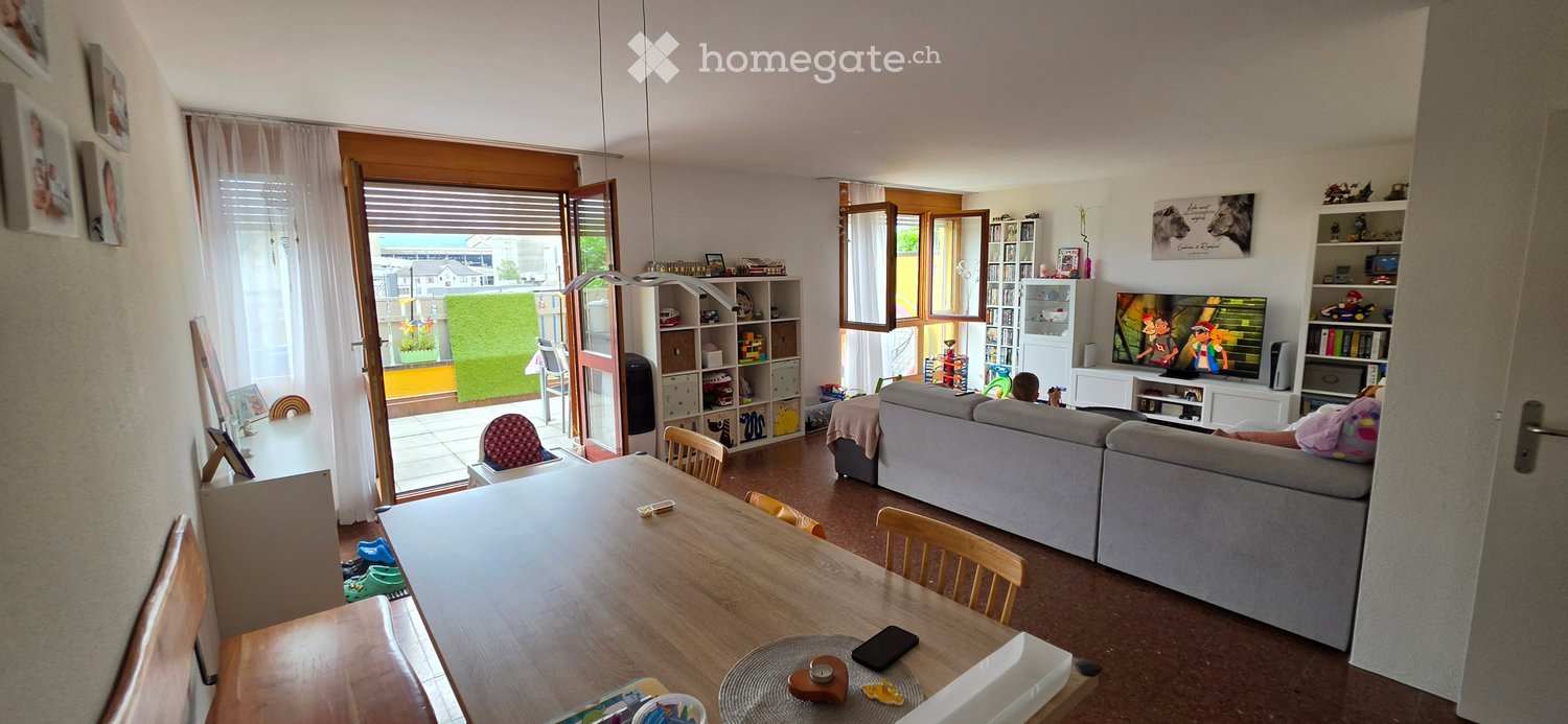 Living room with a grey sofa, wooden dining table, chairs, television, shelves, and picture frames. Large windows and a door leading to a patio.