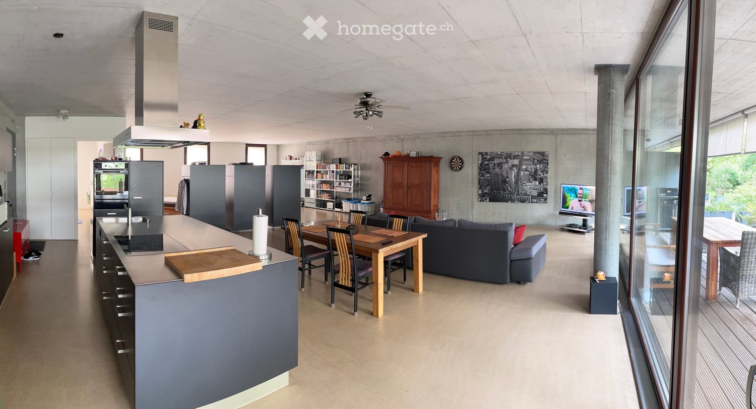Open-plan living and dining area with a modern kitchen, grey sofa, dining table with chairs, and large windows.