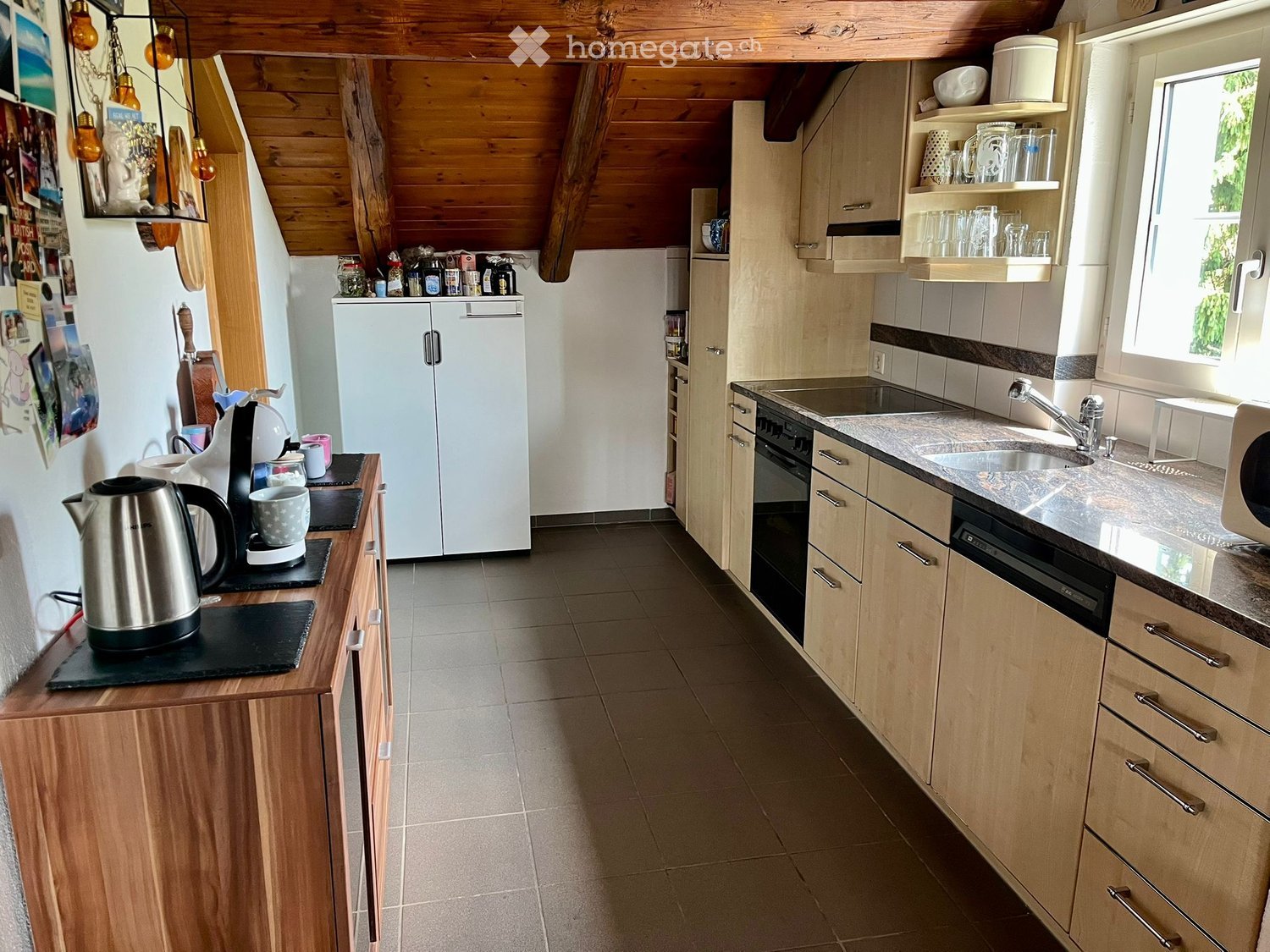 Modern kitchen with light wooden cabinets, stainless steel appliances, and a tiled floor. Features include a refrigerator, oven, dishwasher, sink, microwave, and various kitchen items on the counter.