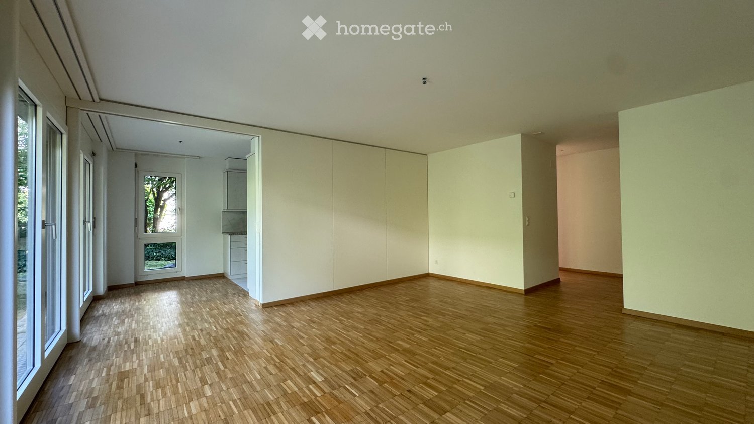 Empty room with wooden flooring, white walls, large windows, and a door leading to another room.