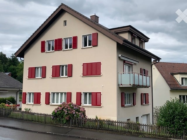 Two-story house with beige walls, red shutters, and a balcony on the upper floor.
