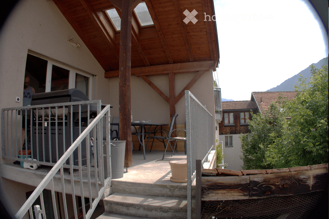 Balcony with wooden roof, metal railing, outdoor furniture including a table and chairs, and a view of mountains in the background.