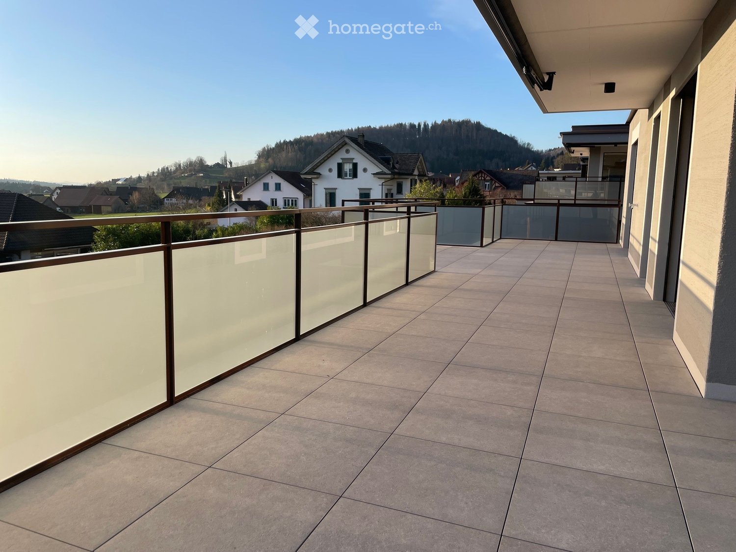 Balcony with tiled floor, glass railing, and a view of a residential area with houses and hills in the background.
