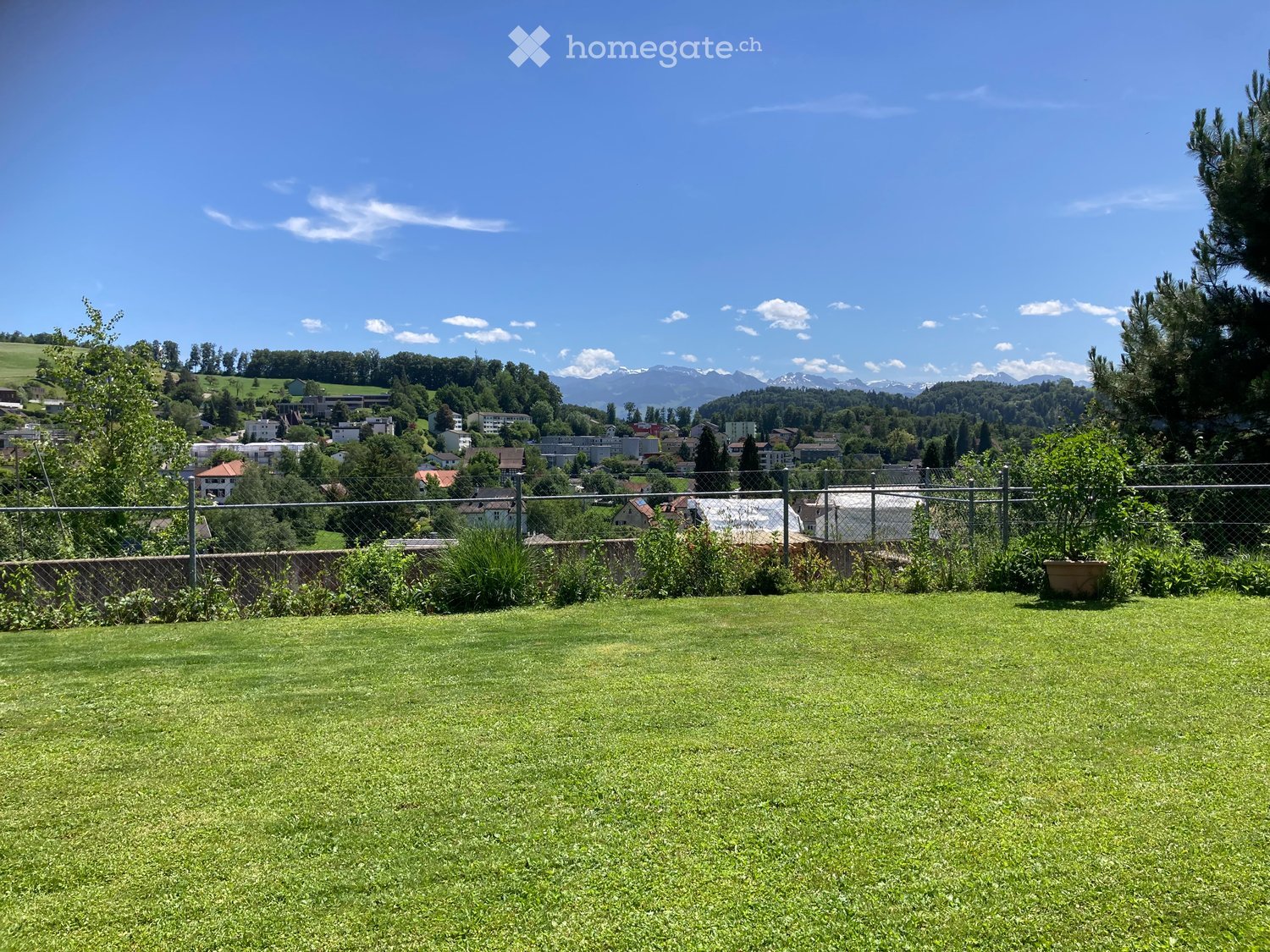 A well-maintained lawn with a metal fence, surrounded by trees and shrubs, overlooking a town with mountains in the background.