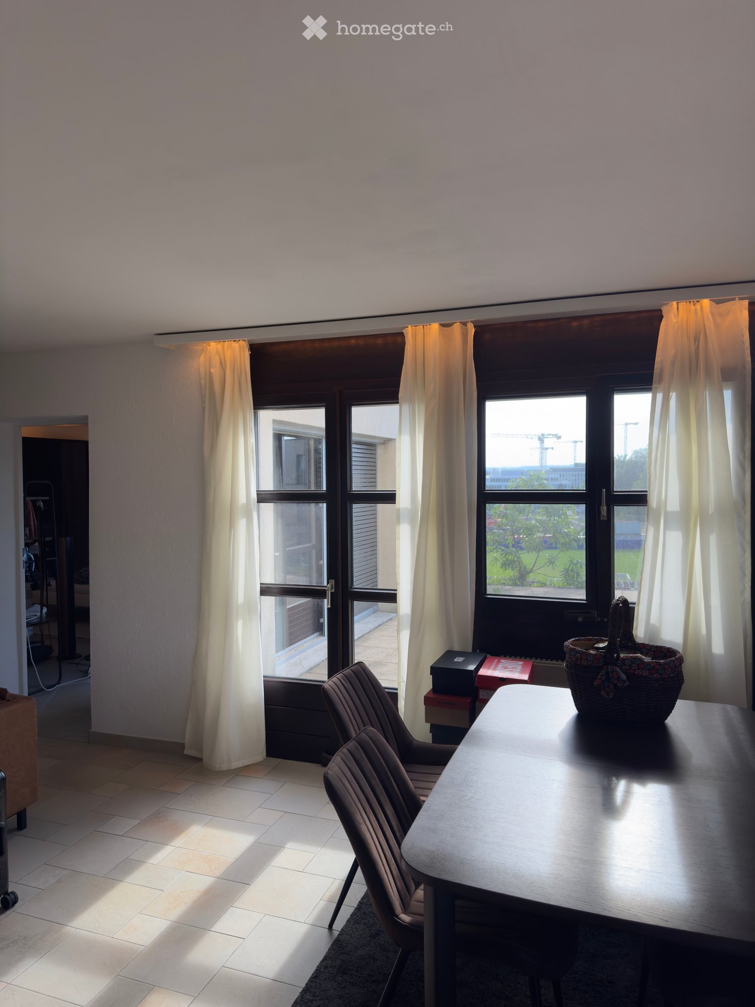 Bright room with large windows and curtains, featuring a wooden table, two chairs, and a decorative basket on the table.