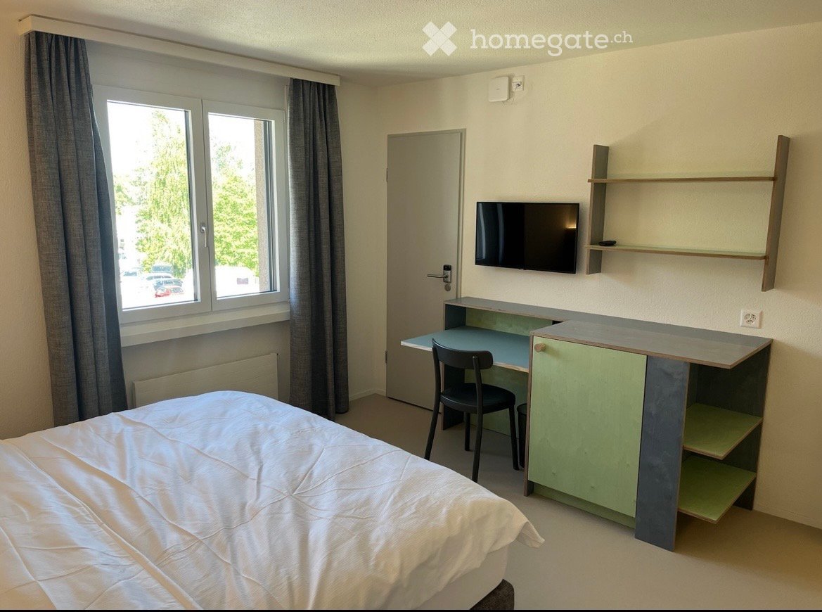 A furnished bedroom with a bed, desk, chair, cabinet, shelves, and a TV mounted on the wall. The room has a window with curtains and a door leading to another space.