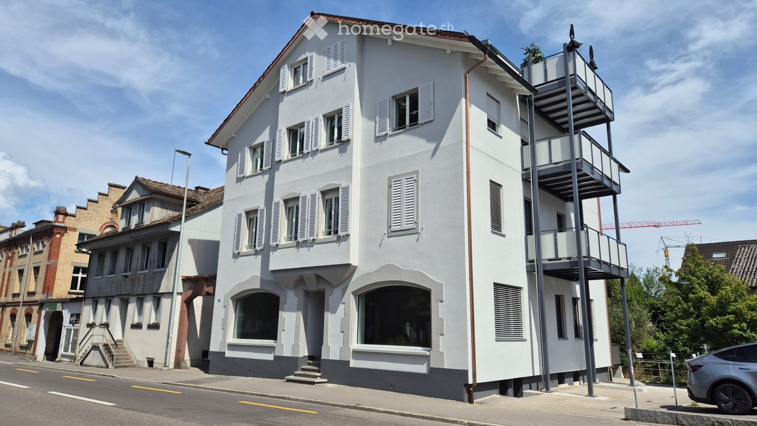 Modern multi-story building with white facade, multiple windows with shutters, and a balcony on the upper level.