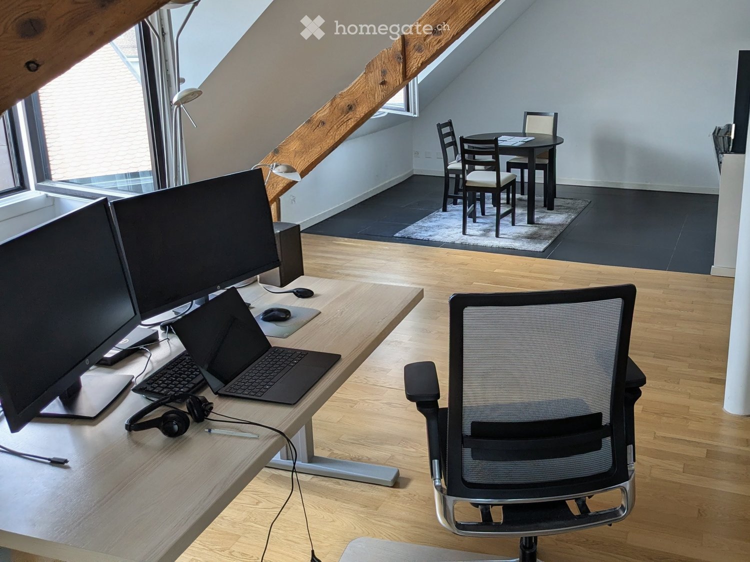 A modern, well-lit room with wooden flooring, a large desk with a laptop, dual monitors, and a comfortable office chair. A small dining area with a table and chairs is visible in the background.