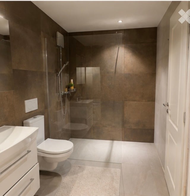 Modern bathroom with dark brown tiles, white fixtures, glass-enclosed shower, and a small shelf holding toiletries.