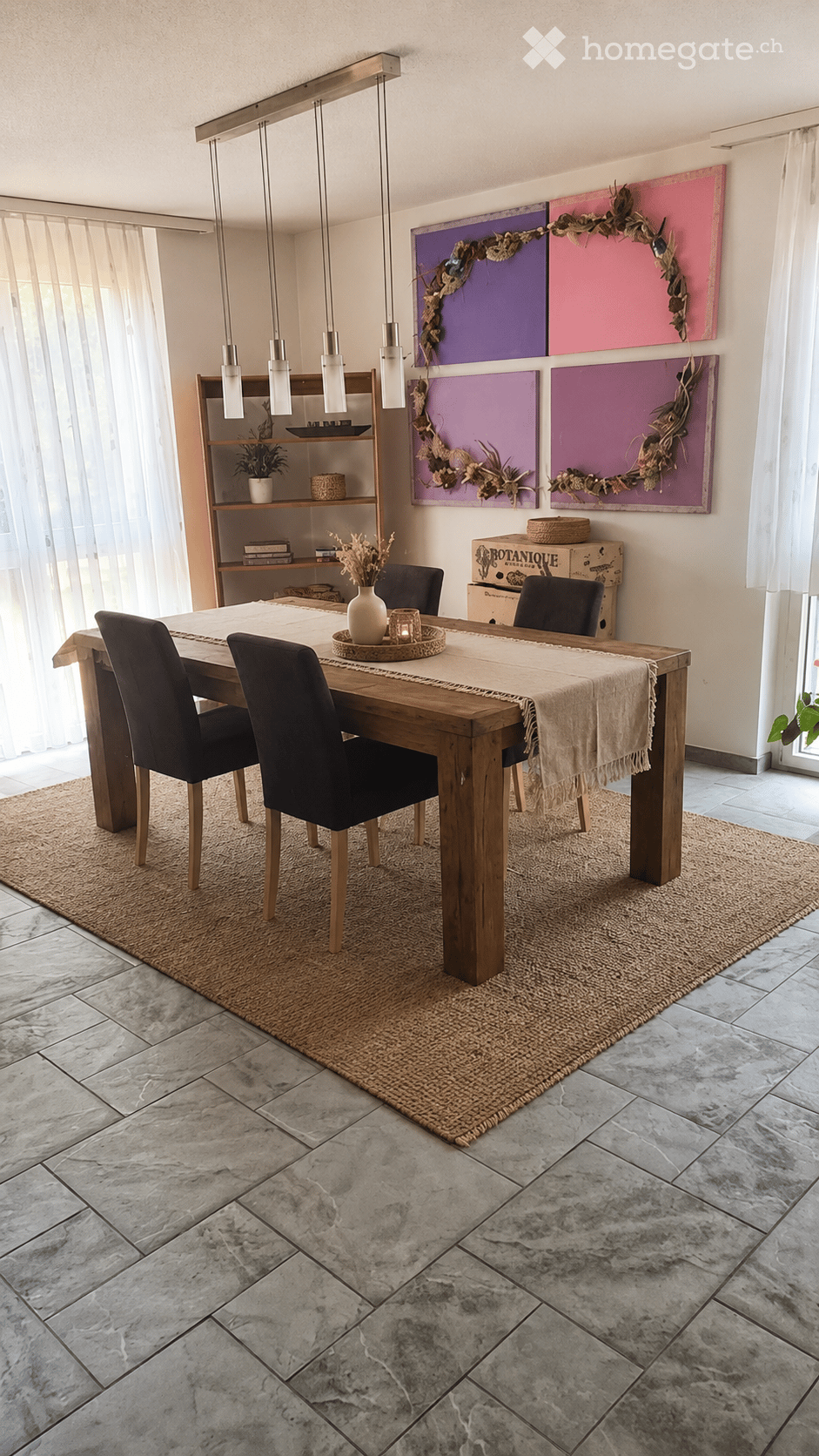 Dining room with a wooden dining table, four black chairs, a beige table runner, a white vase with dried flowers, a shelf with books and a small plant, and colorful wall art.