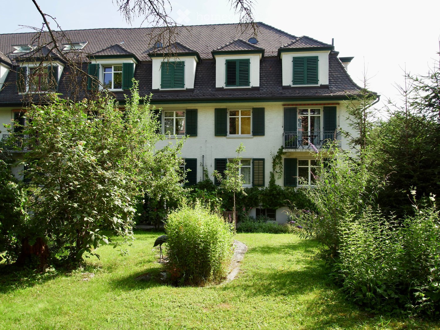 Two-story house with white exterior, green shutters, multiple windows, and a well-maintained garden with a pathway leading to the entrance.