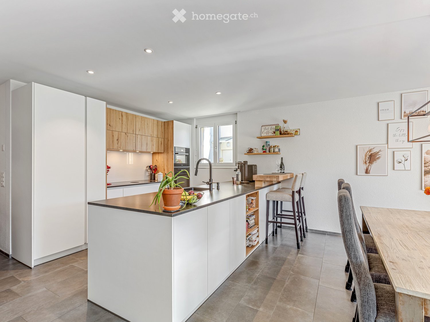 Modern kitchen with white and wooden cabinets, island, bar stools, dining table, and various decorative items on shelves and walls.