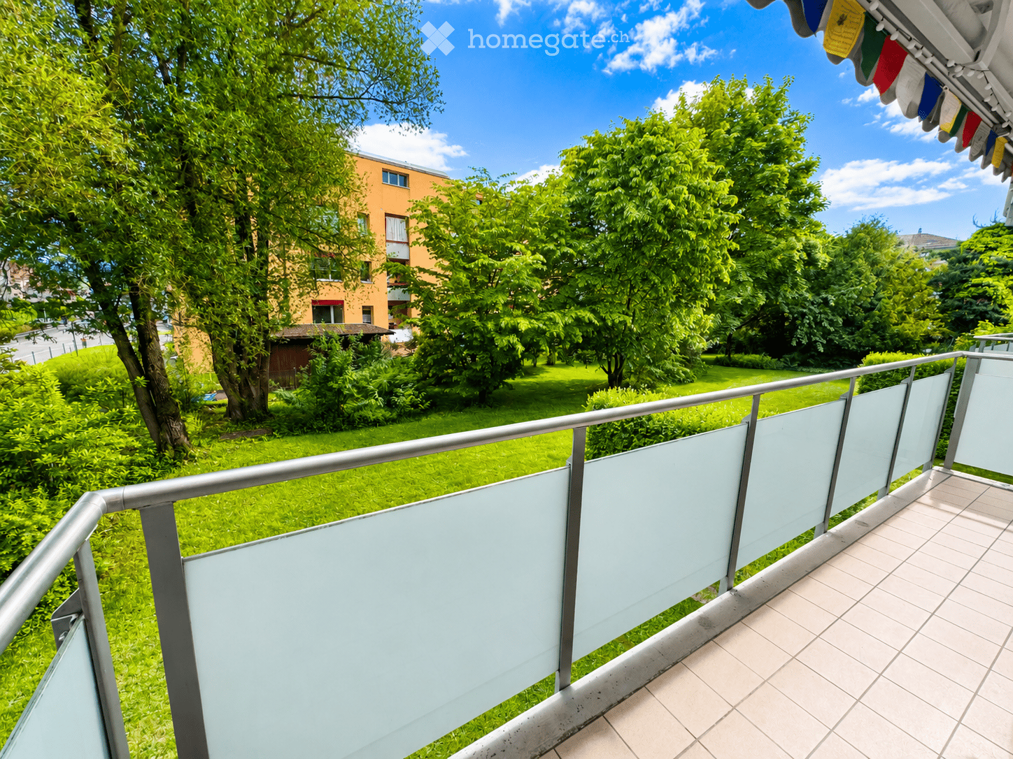 Balcony with modern glass railing, tiled floor, overlooking a green lawn and trees.