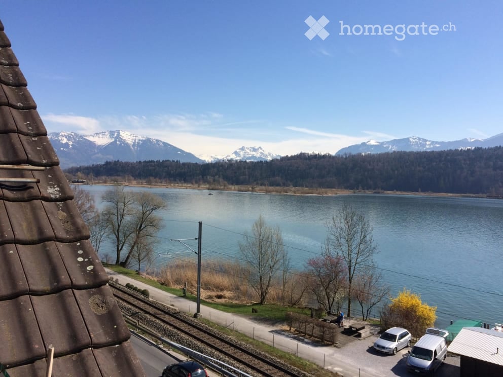 View from a rooftop showing a serene lake surrounded by snow-capped mountains, with a road running alongside and parked cars visible.